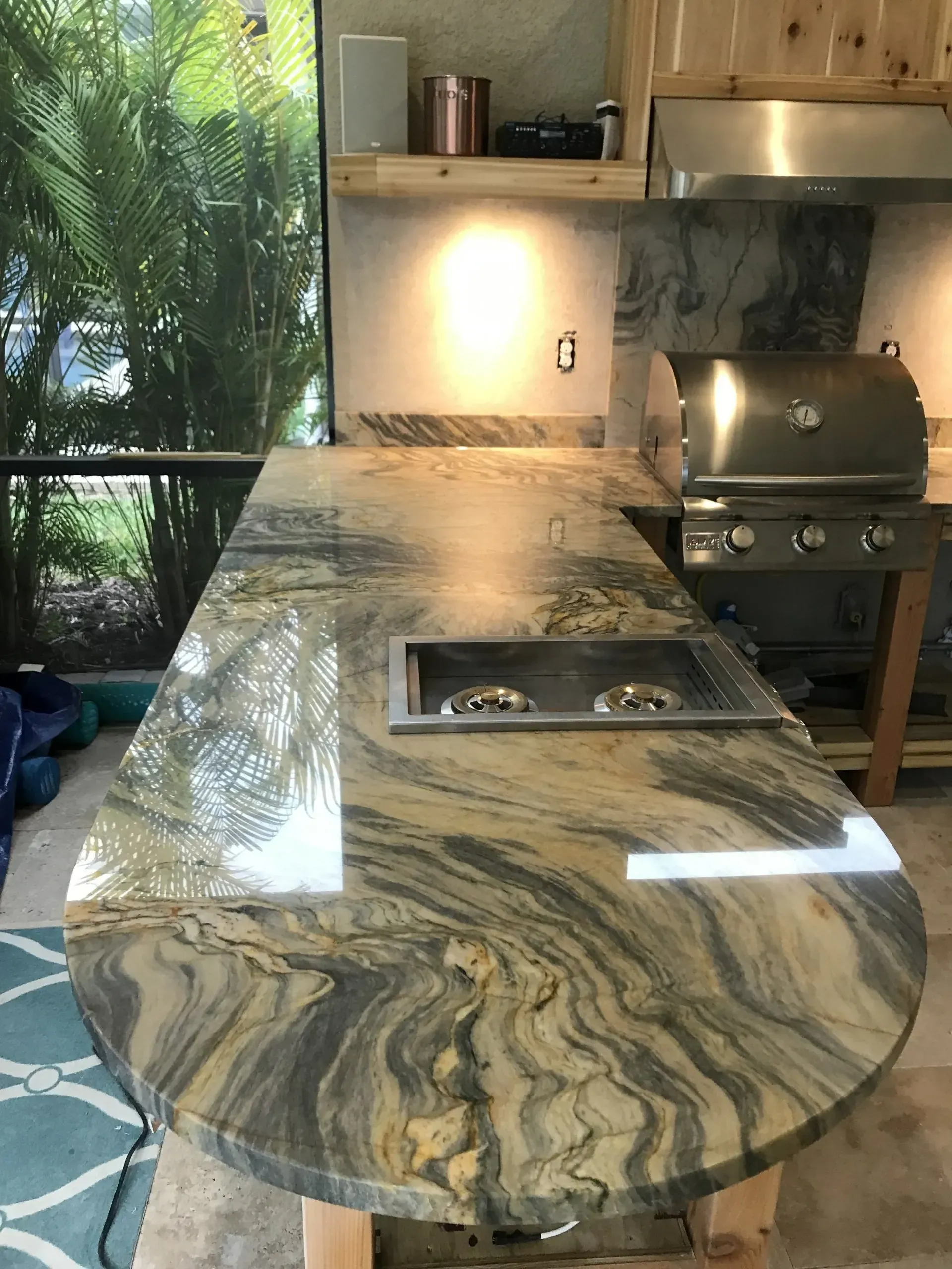Polished granite outdoor kitchen island with built-in grill and sink, beside a patio and lush greenery