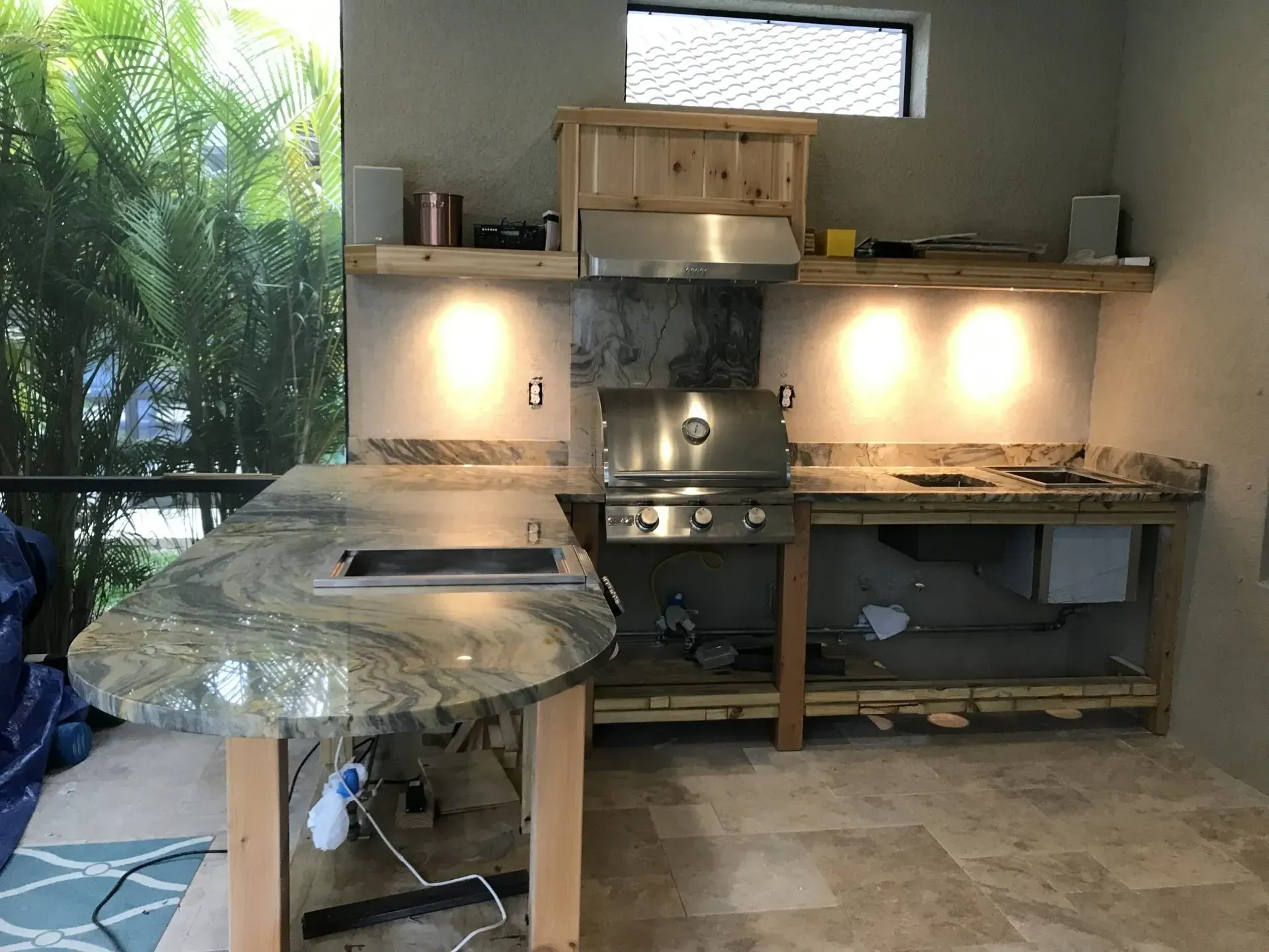 Outdoor kitchen with granite counters, stainless grill, wood cabinets, and a round table covered in plastic.
