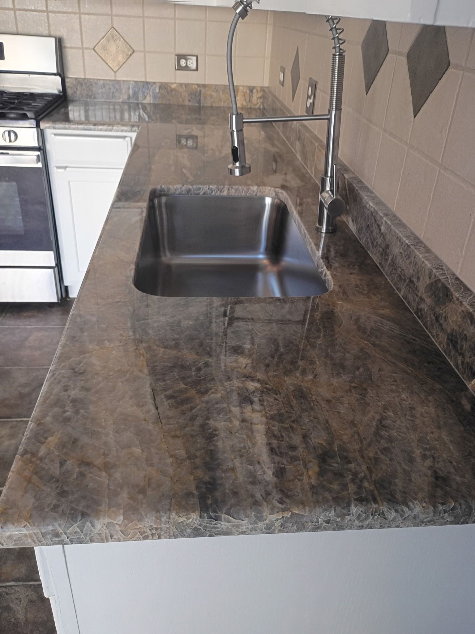 Kitchen counter with a stainless steel sink and dark stone countertop beside a stove.