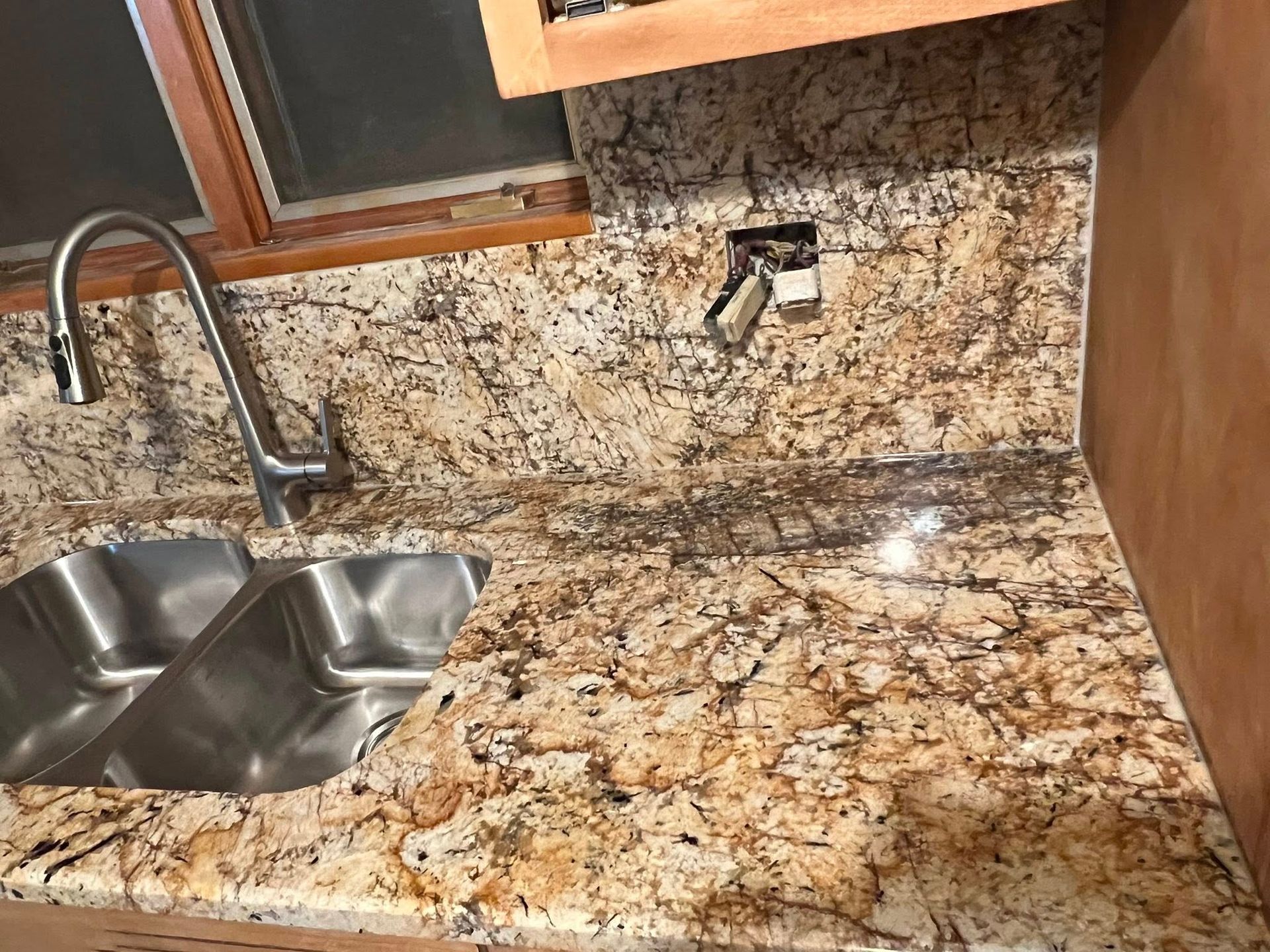 Kitchen countertop with speckled granite, double sink, faucet, and wall outlet under wooden cabinets