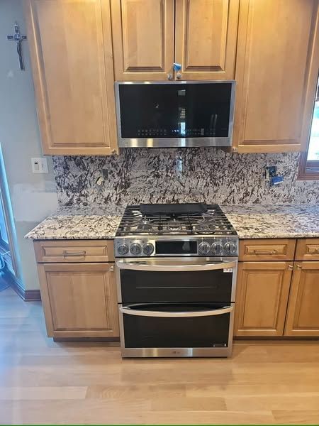 Kitchen with stainless steel stove and microwave beneath wood cabinets and granite backsplash.
