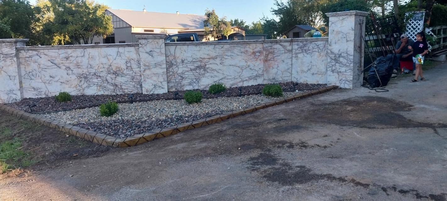 Empty paved lot beside a white stone wall and a fenced gate, with a small landscaped bed.