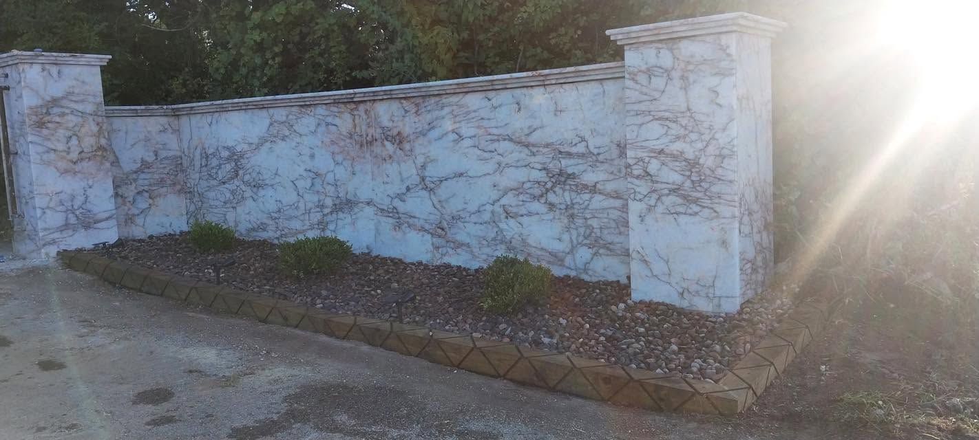 White stucco wall with blue trim beside a roadside, covered in vine shadows and bright sunlight.
