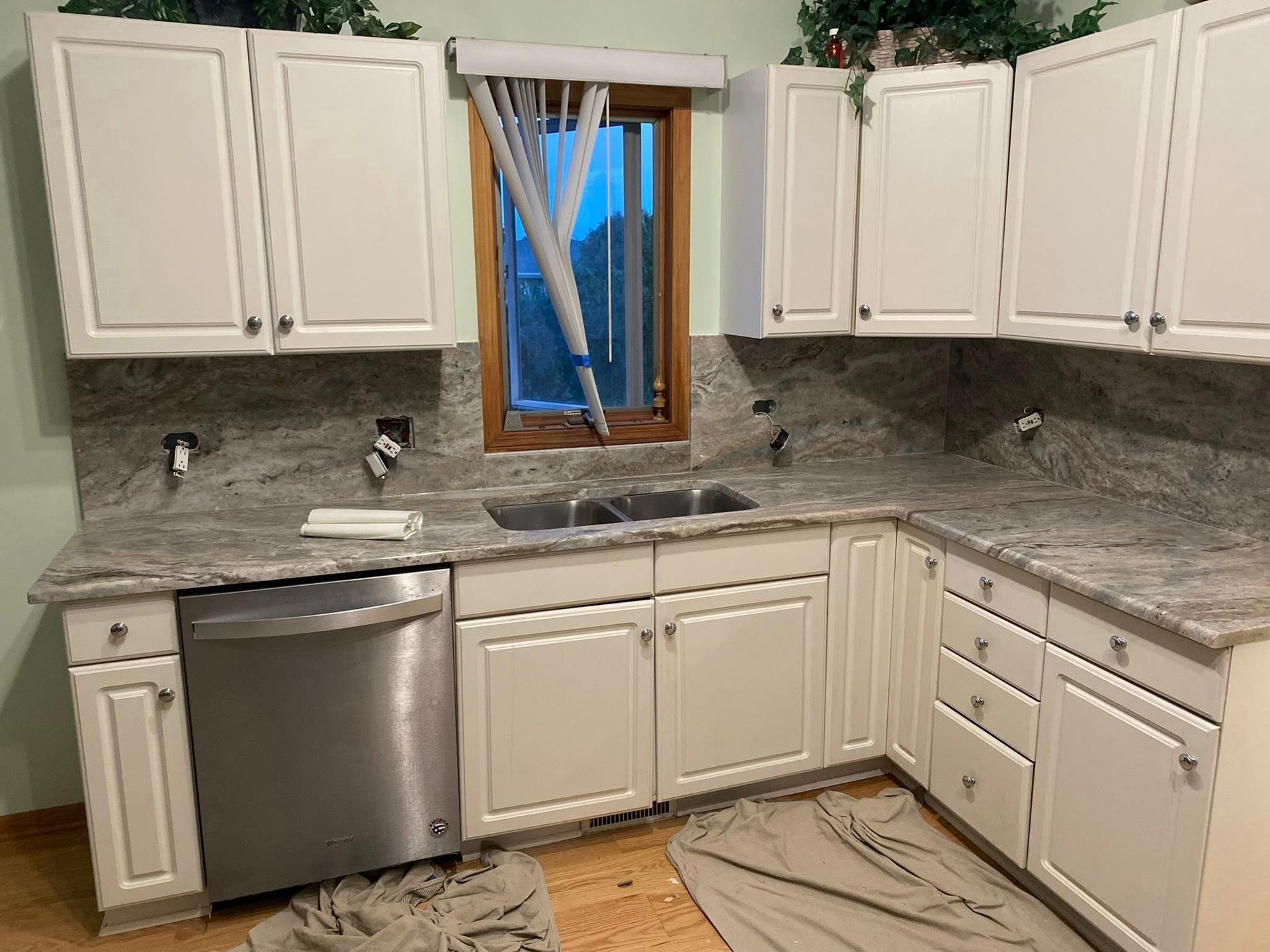 White kitchen with granite counters, stainless dishwasher, window above sink, and unfinished floor repair