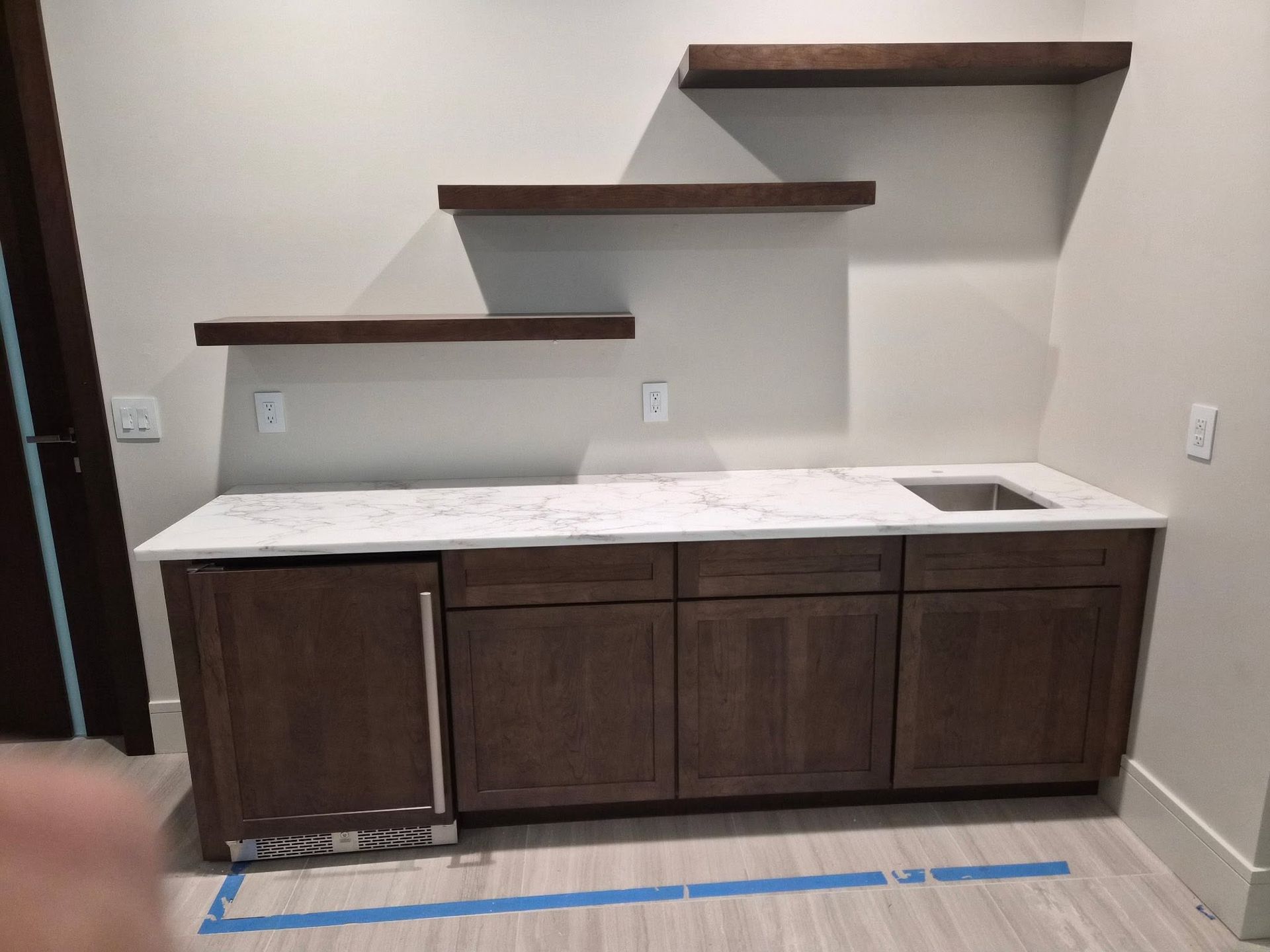 Small kitchen nook with dark cabinets, white countertop, sink, and two floating shelves above it.