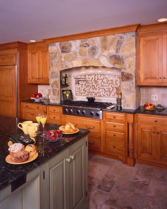 Warm wood kitchen with stone backsplash, gas range, and center island topped with fruit and bread.