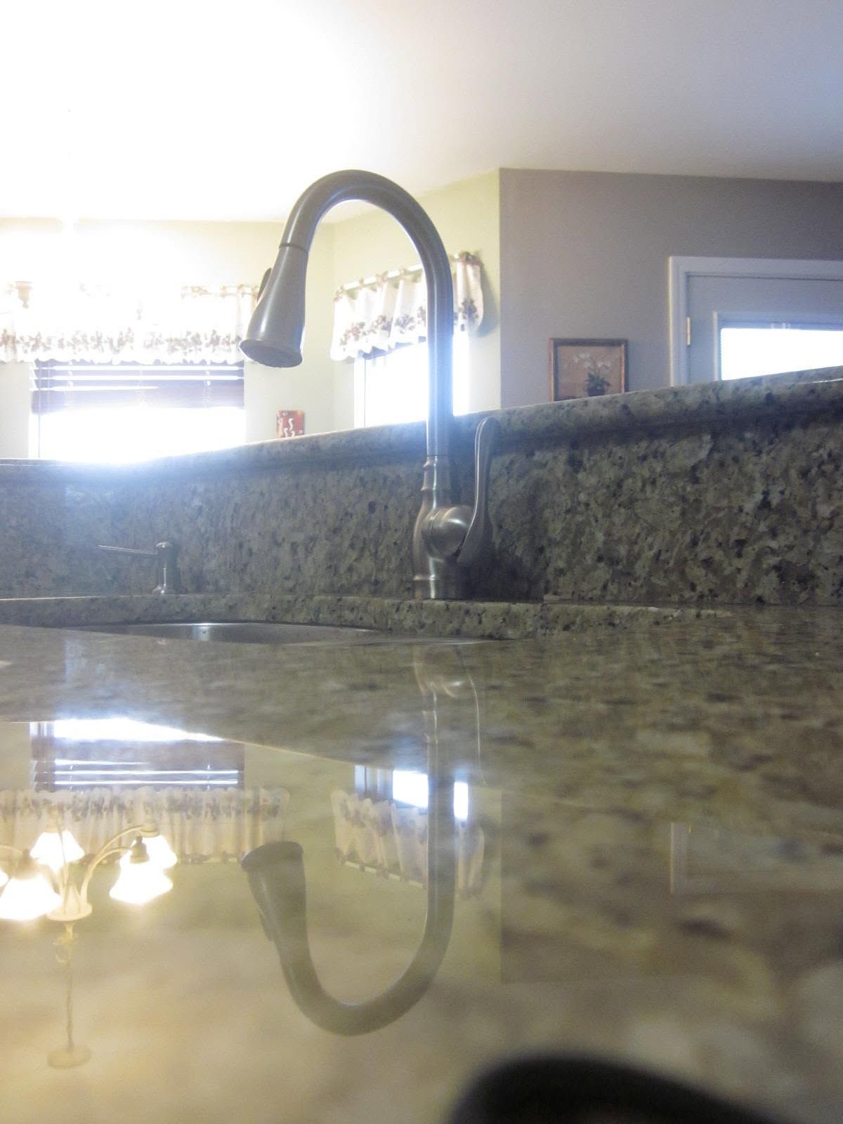 Kitchen sink faucet reflected in a glossy granite countertop, with a bright kitchen in the background.