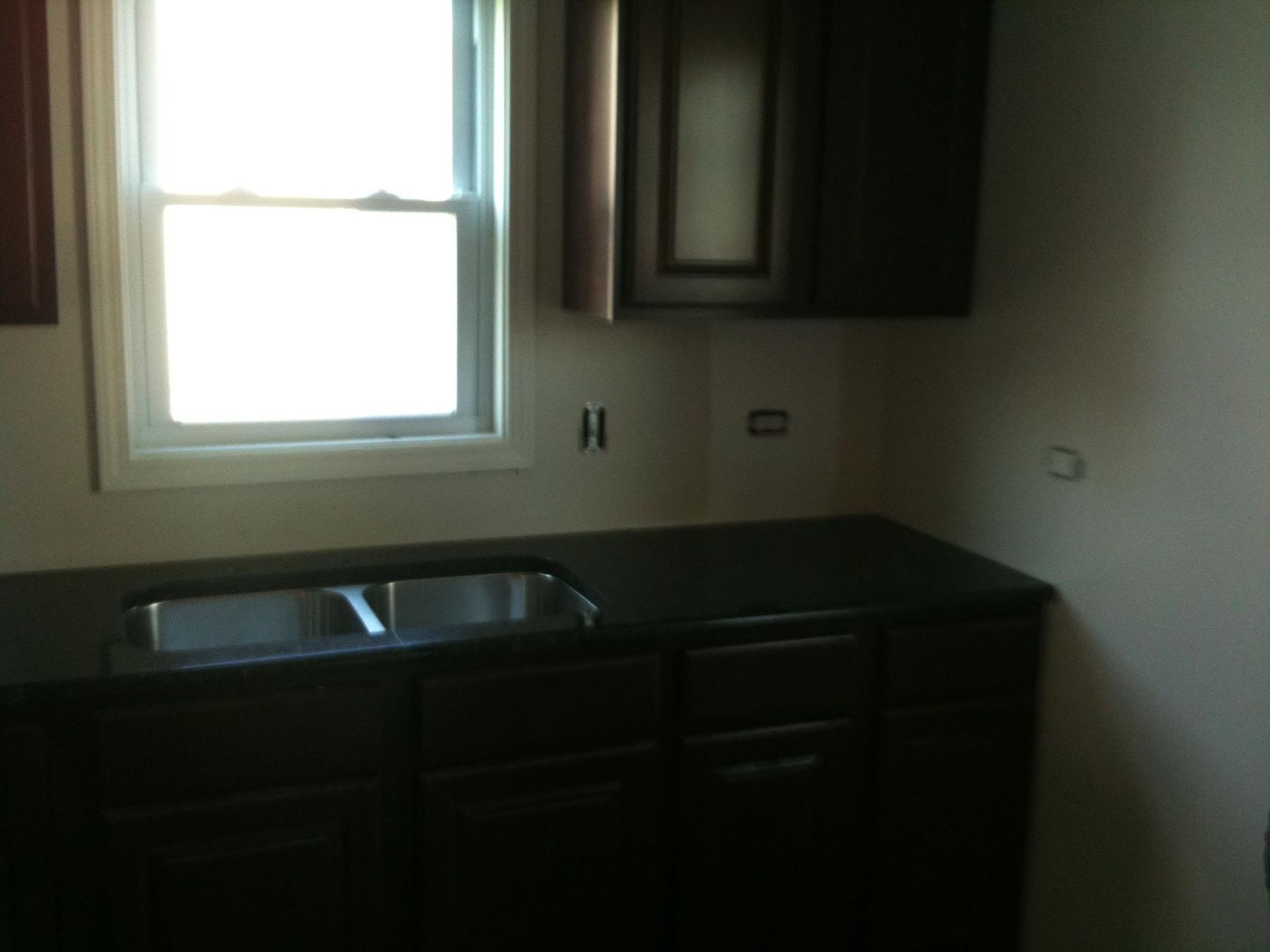 Empty kitchen with dark cabinets, black countertop, sink under a bright window.