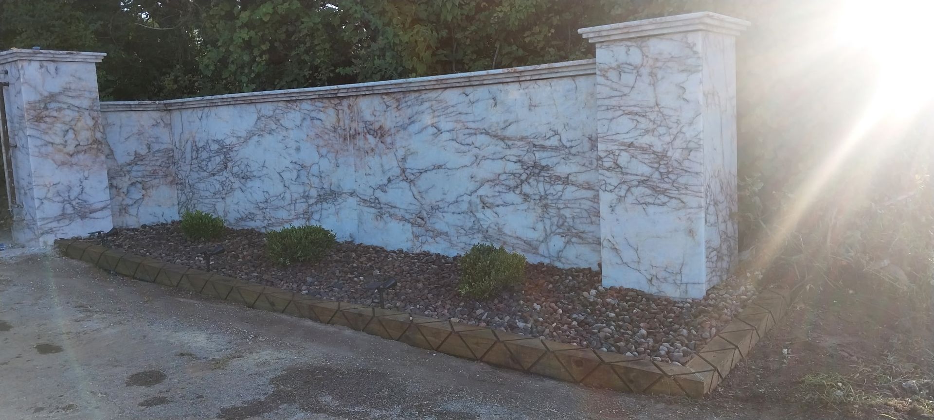 White stucco wall with blue graffiti in sunlight beside a gravel path and trees
