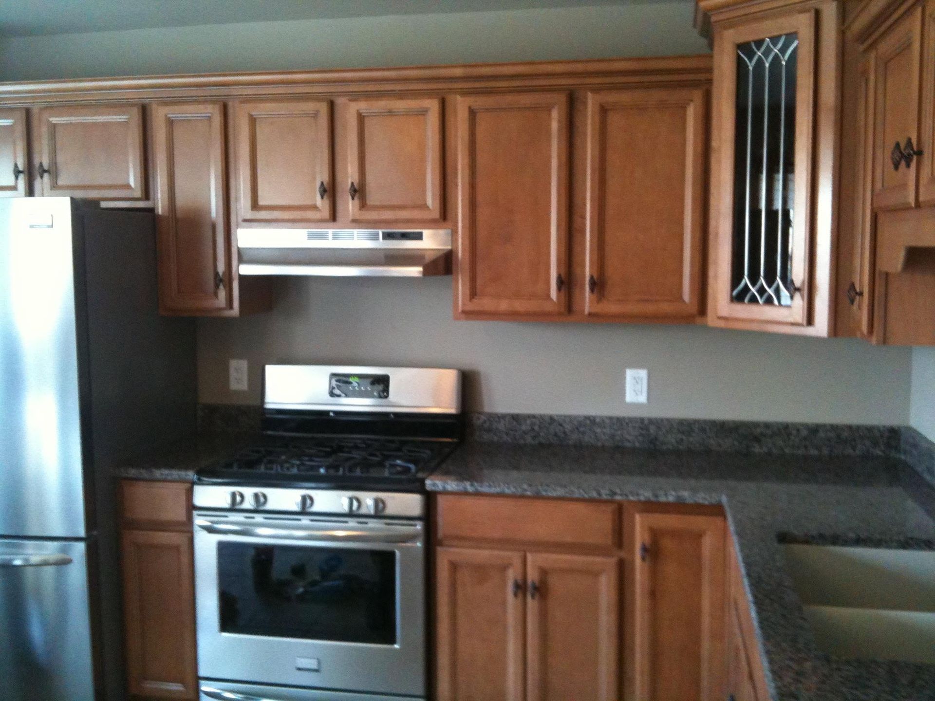 Kitchen with wood cabinets, stainless stove and fridge, dark granite countertop, and glass-front cabinet.