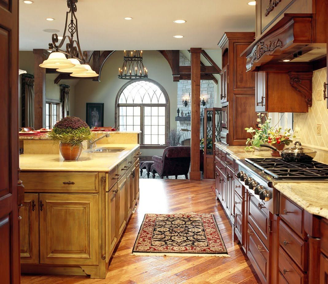 Warm kitchen with wood cabinets, marble island, hanging lights, and a rug leading to a bright arched window.