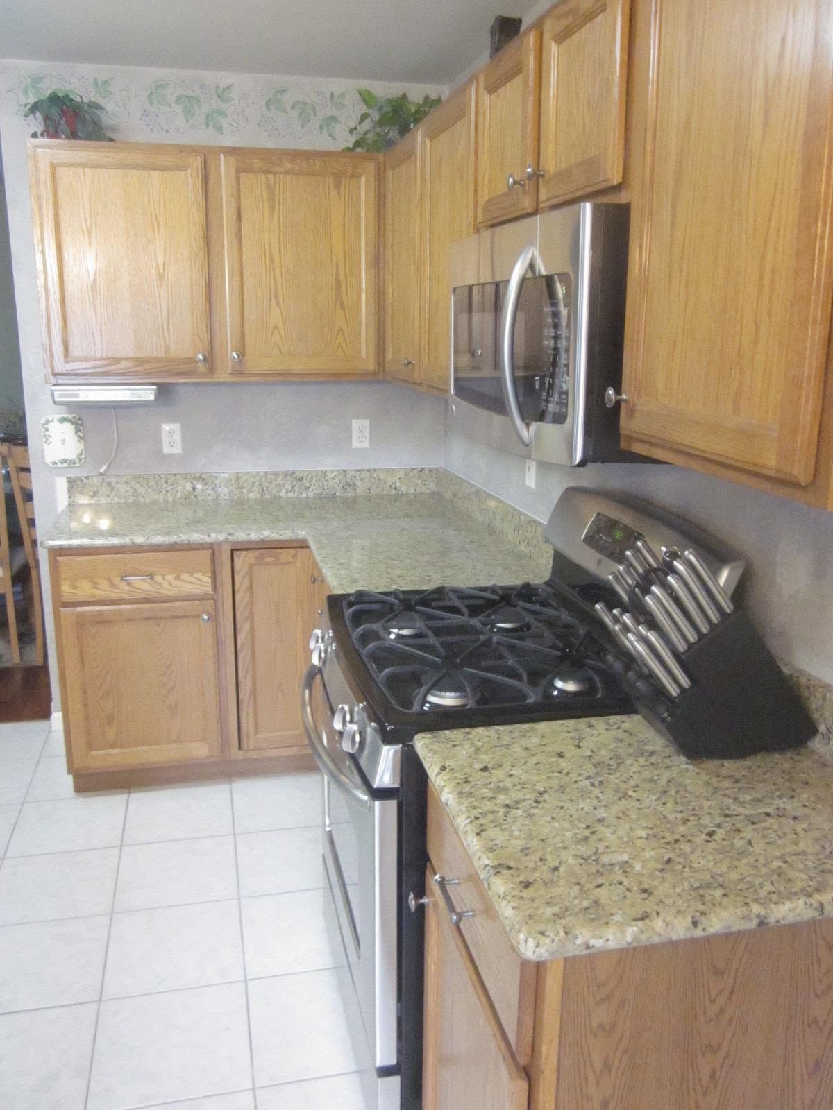 Kitchen with light wood cabinets, speckled countertop, white tile floor, and stainless steel stove and microwave.