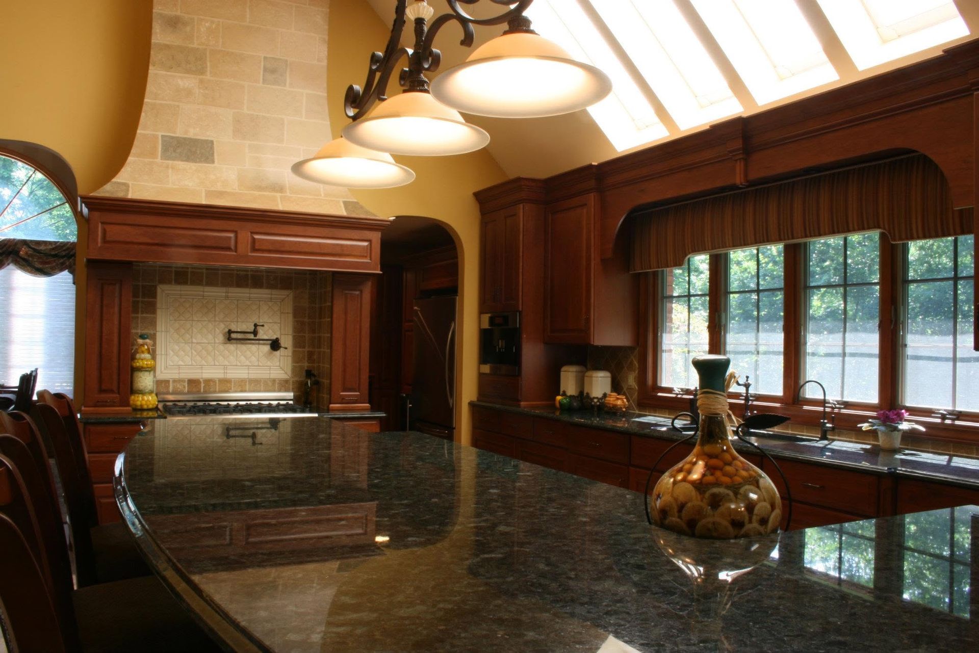 Luxurious kitchen with dark granite island, wood cabinets, pendant lights, and large windows overlooking greenery