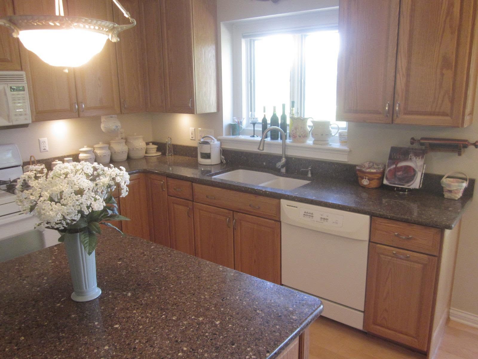 Kitchen with wood cabinets, black countertops, sink under window, and vase of white flowers on island.