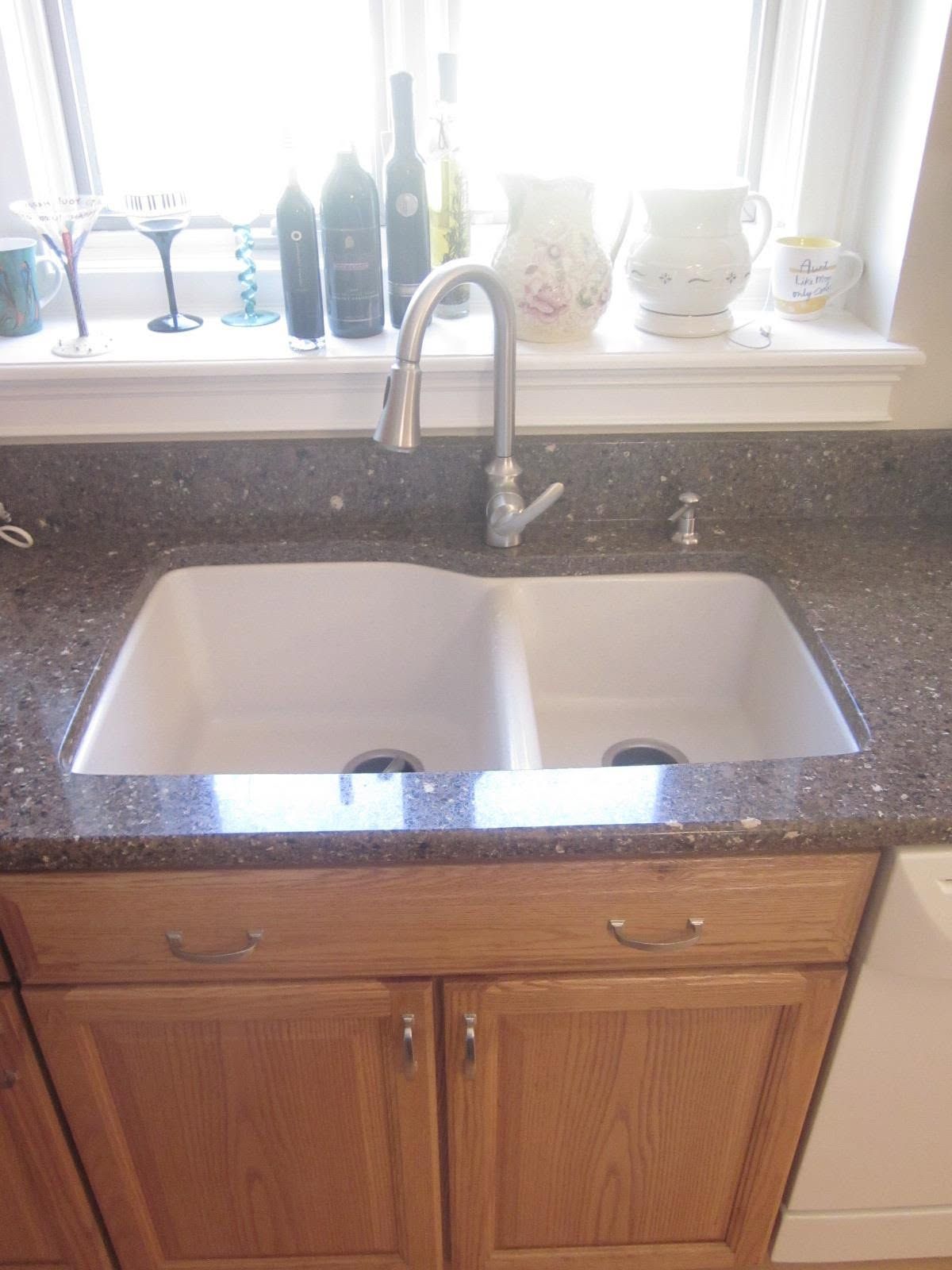 Double kitchen sink with chrome faucet on a speckled countertop beneath a window with bottles and decor.