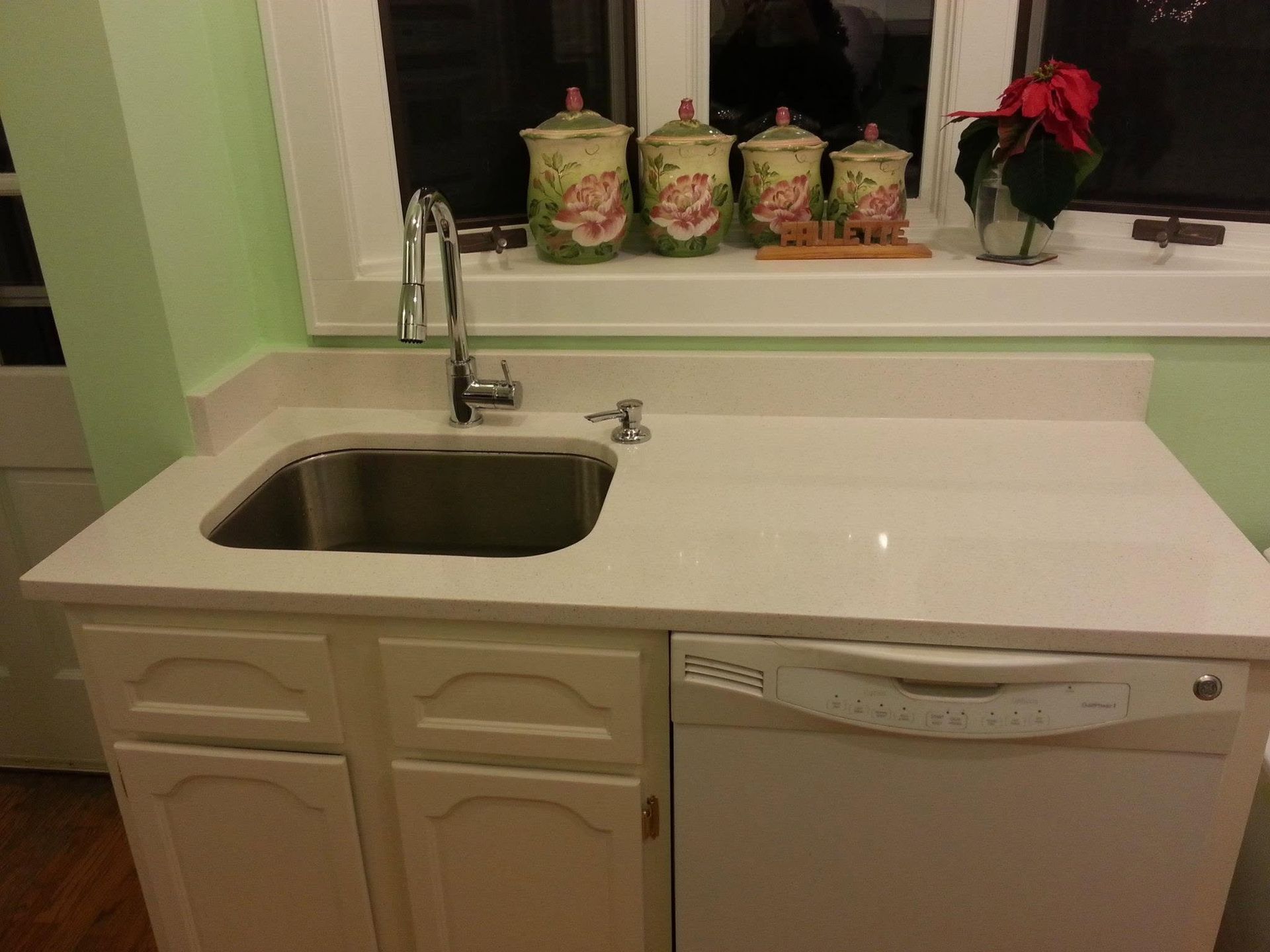 Kitchen sink and countertop with soap dispensers, a dishwasher, and a window with red flowers.