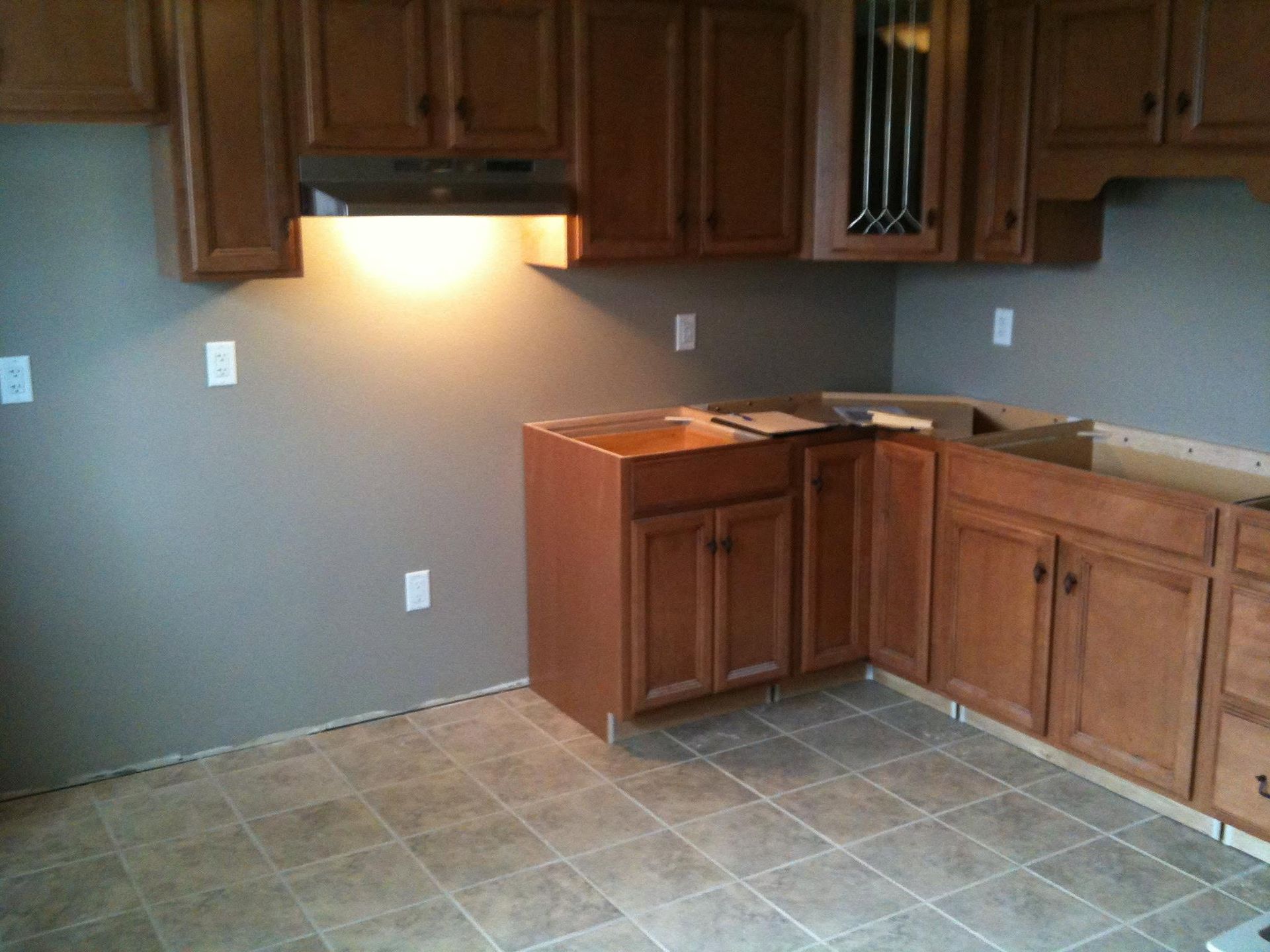 Empty kitchen with brown cabinets, gray walls, and beige tile floor under a lit stove hood
