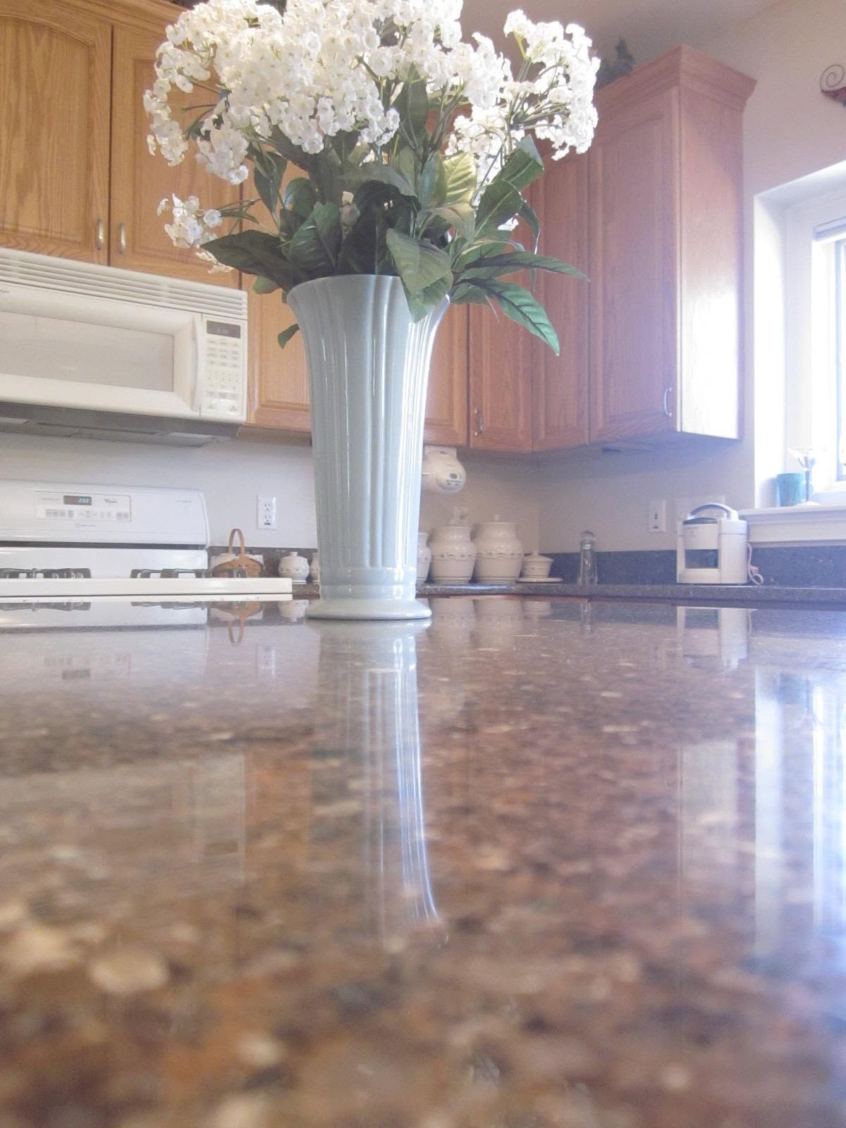 White flowers in a tall vase on a kitchen countertop with wooden cabinets and bright window light.