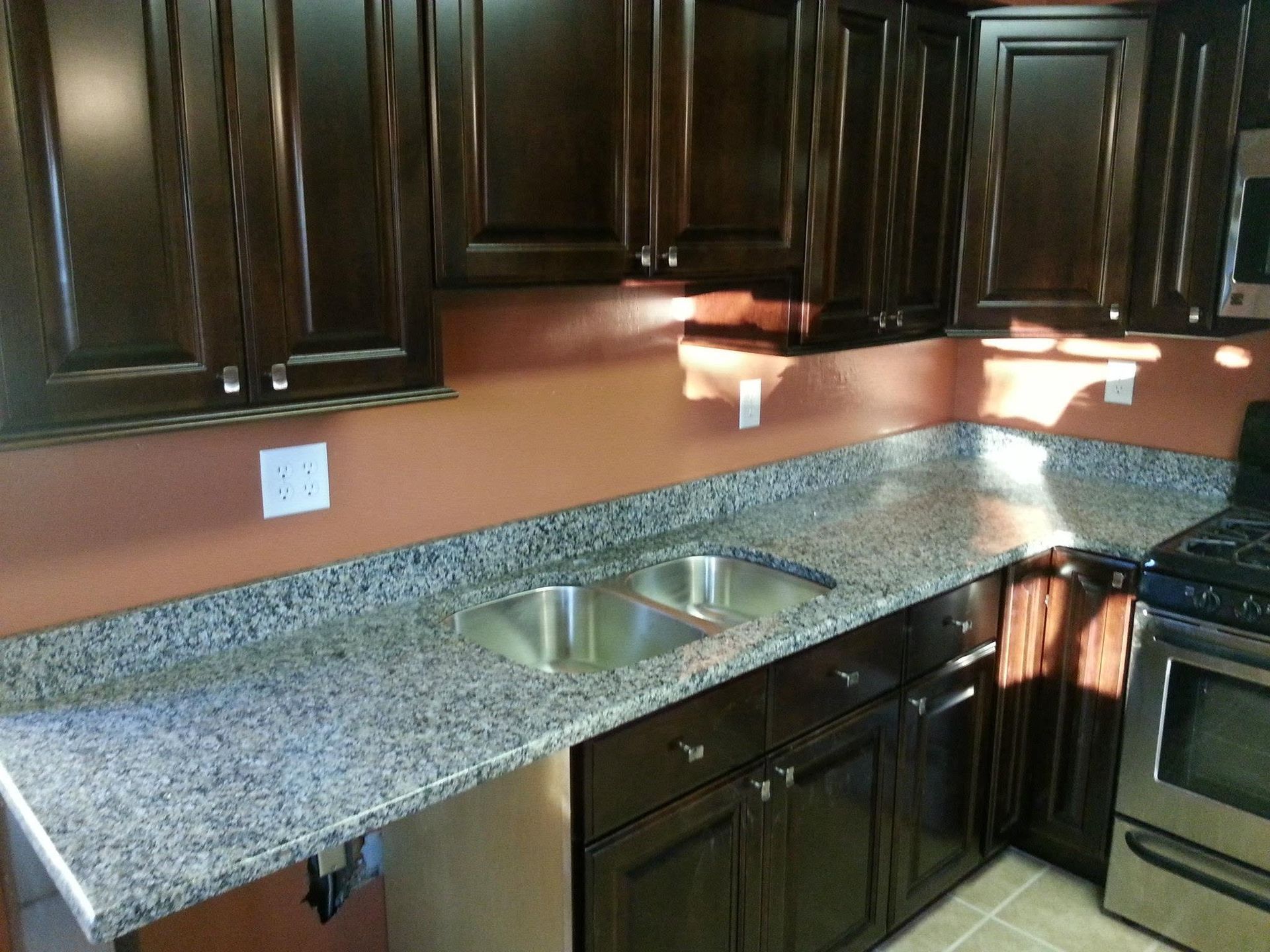 Granite kitchen countertop with double sink, dark wood cabinets, and stove on the right.