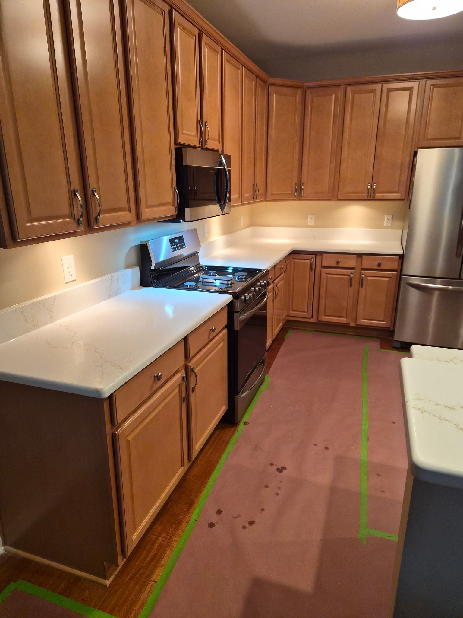 Empty kitchen with wood cabinets, white countertops, stainless fridge, and tiled floor.