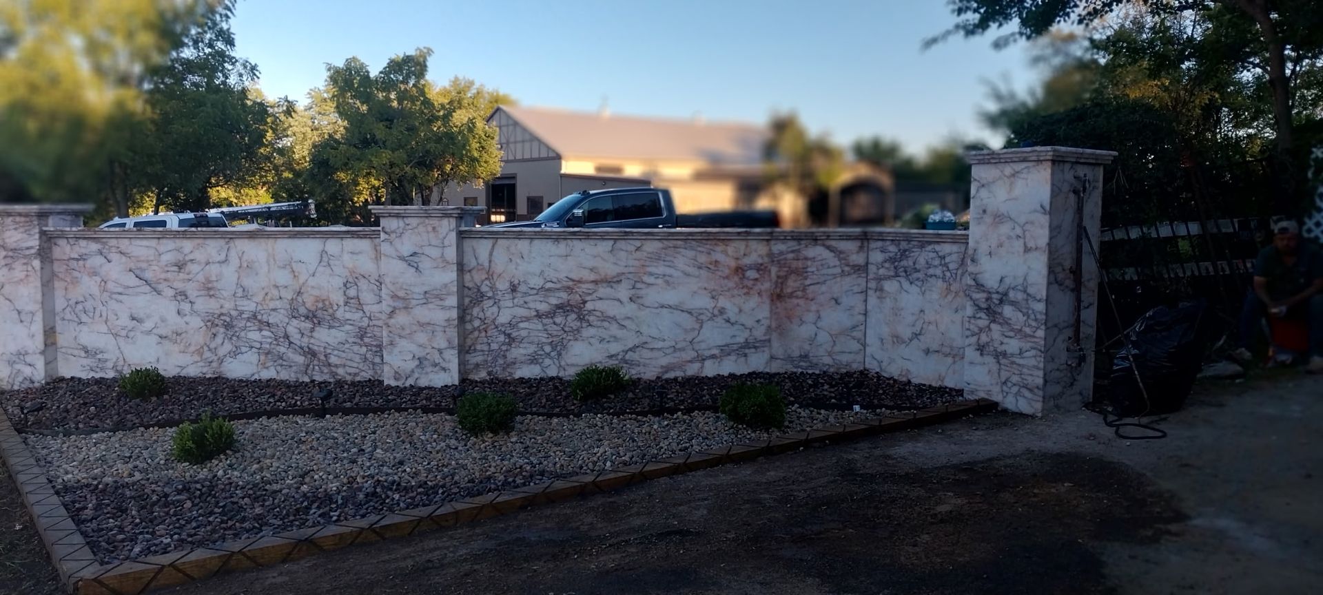 Sunlit house behind a white wall and gate, with a parked blue car and gravel driveway.