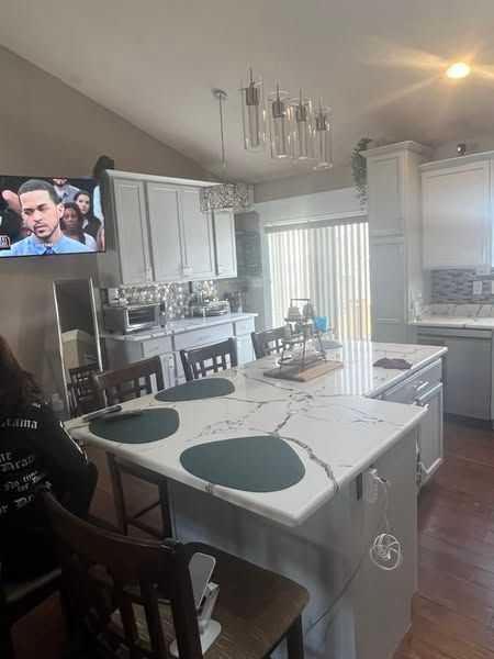 Open kitchen with white cabinets, island, dining chairs, and a TV showing a man in the corner.