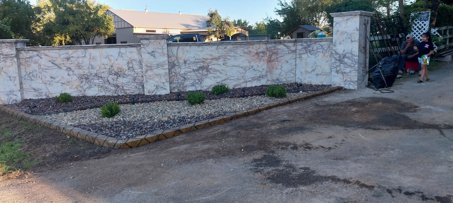 Stone wall beside a dirt driveway, with a small gravel bed and trees in the background