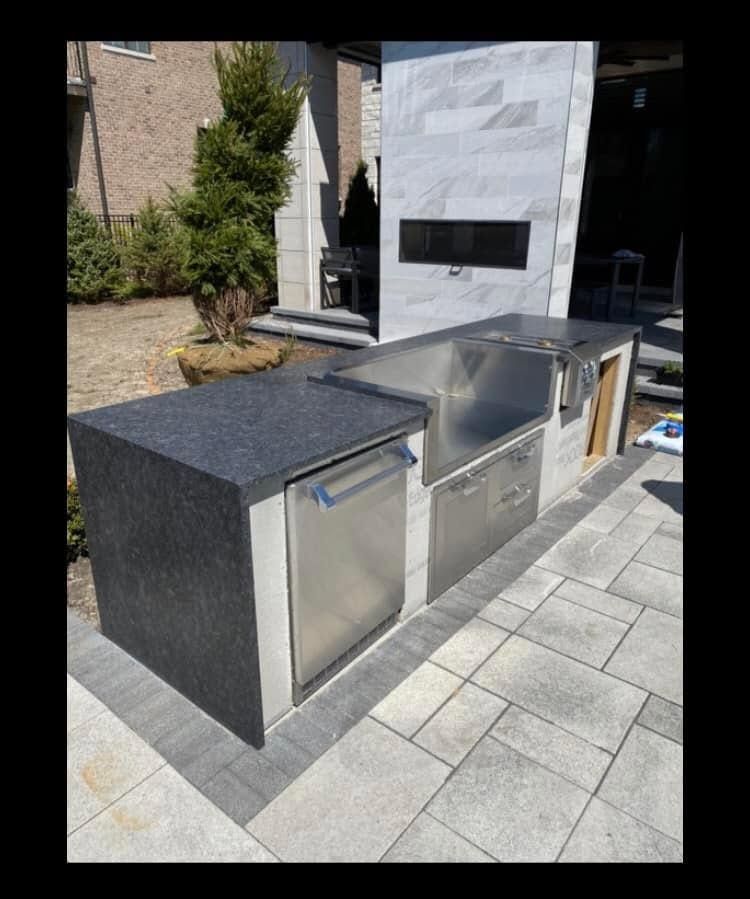 Outdoor stainless-steel grill and sink island with black countertop on a stone patio beside a white fireplace.