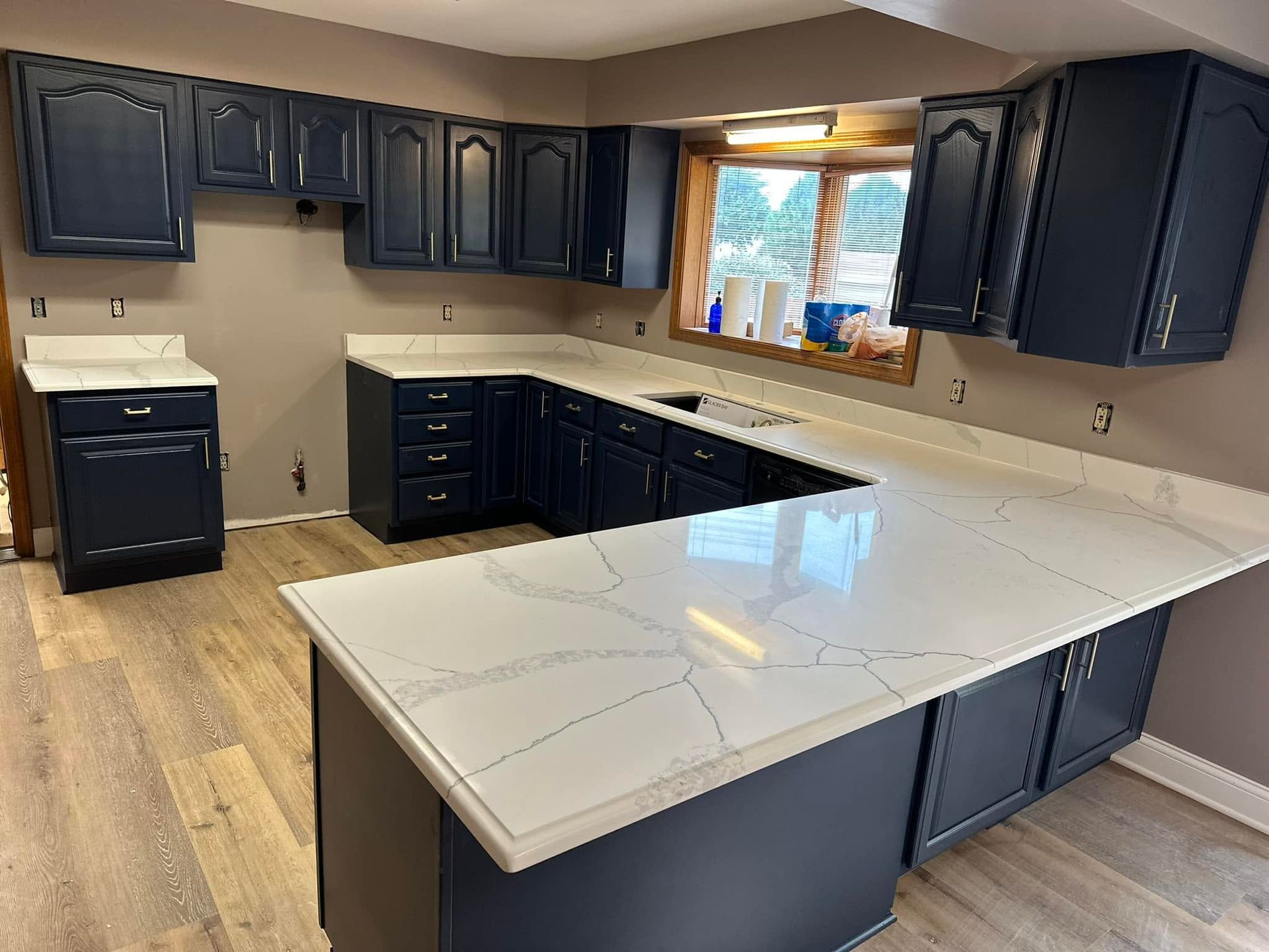 Empty kitchen with dark cabinets, white countertop island, beige walls, and a window over the sink.