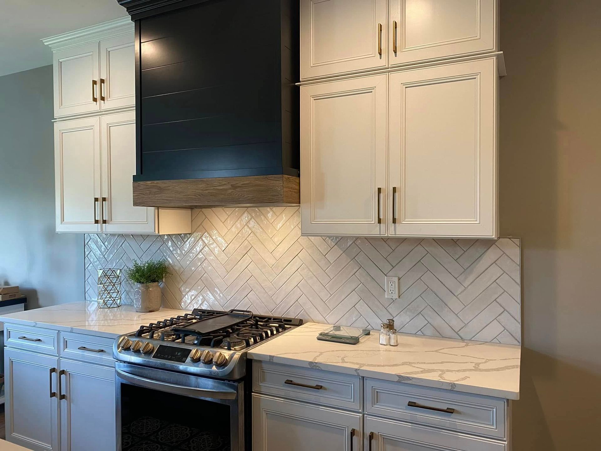 Modern kitchen with white cabinets, black range hood, herringbone backsplash, and stainless steel stove.