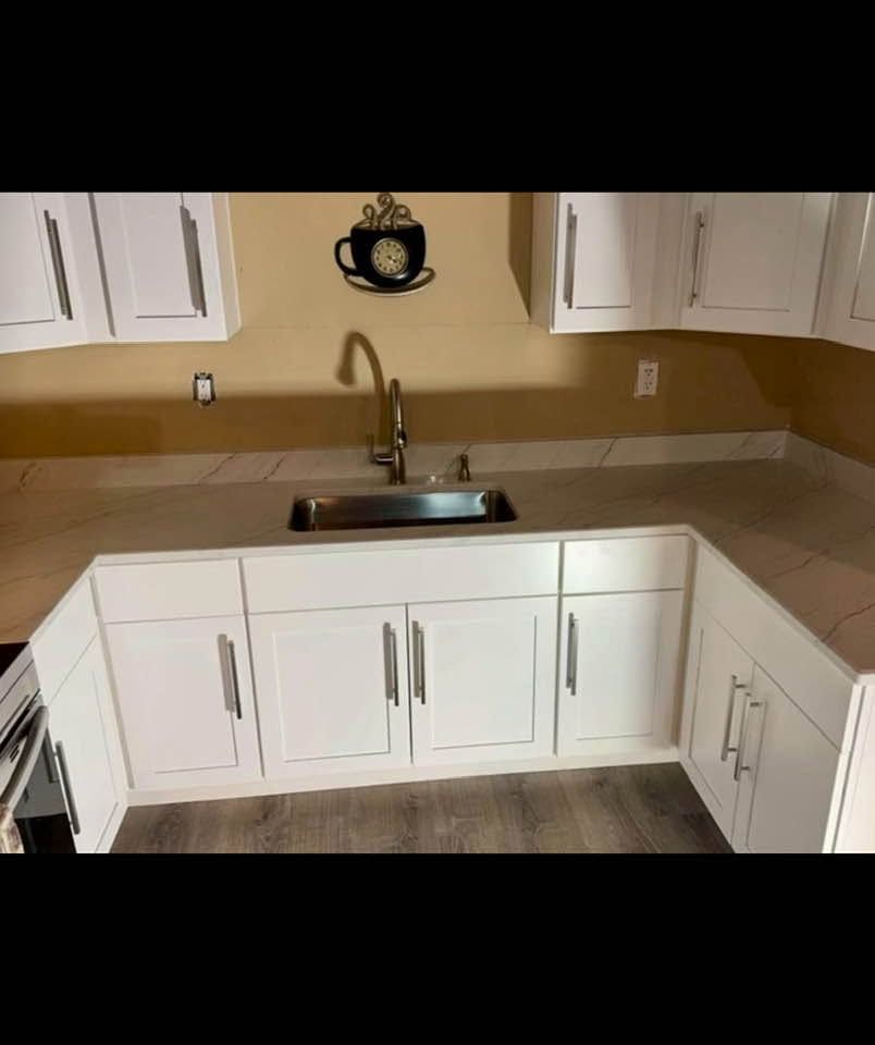 White kitchen corner with sink, tan countertop, and cabinets under a wall-mounted clock-like decoration