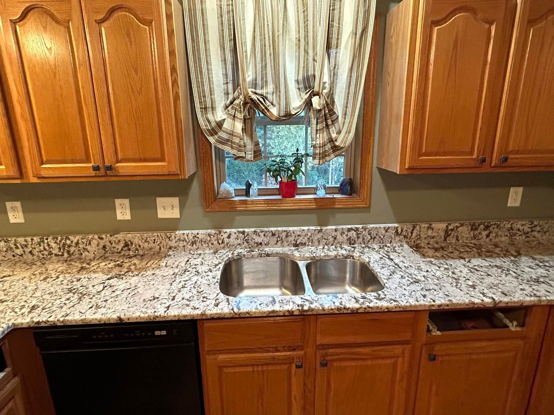 Kitchen counter with speckled granite, double sink, wooden cabinets, and a window with curtains above the sink