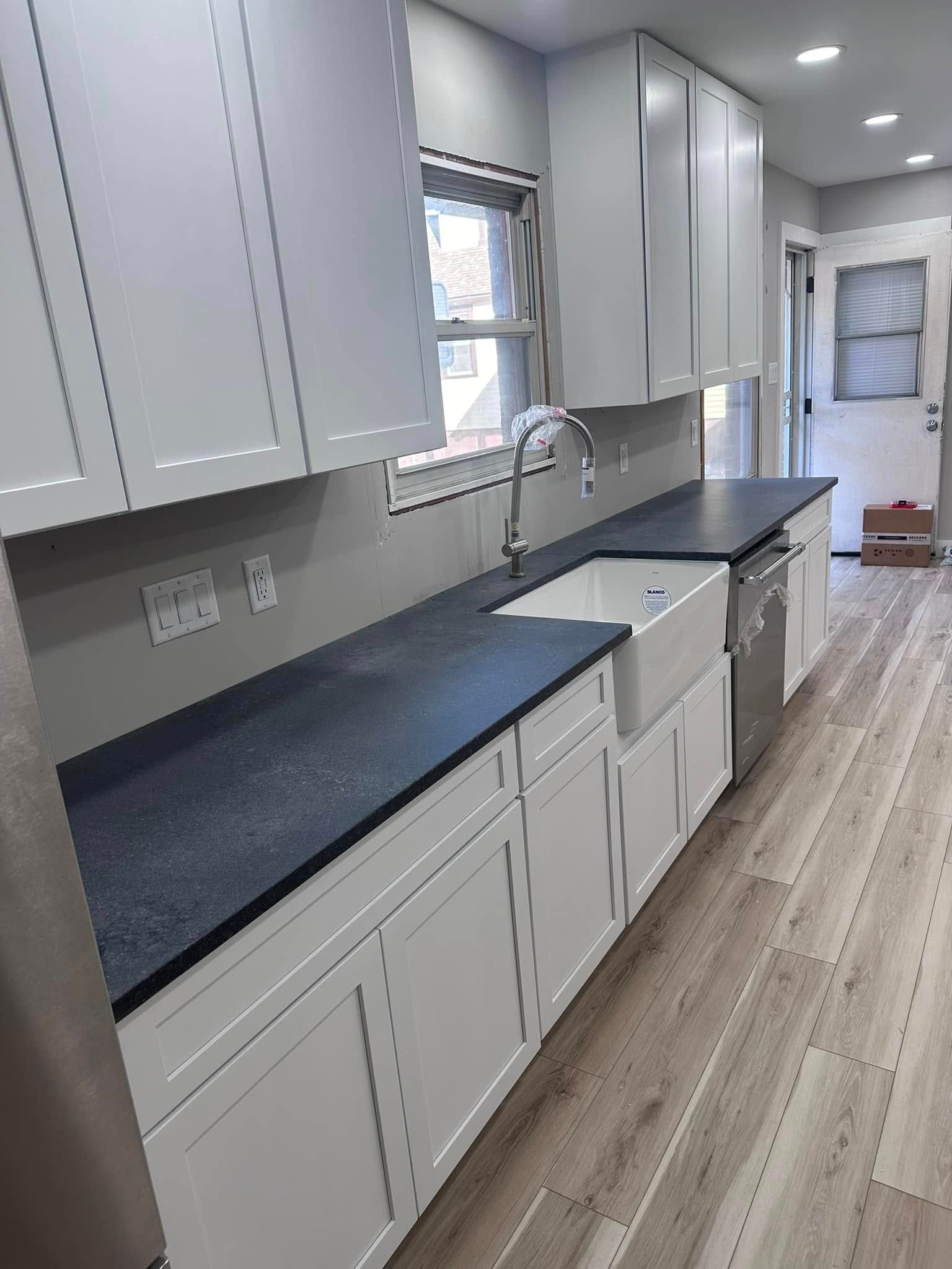 Bright galley kitchen with white cabinets, dark countertops, stainless sink, and light wood flooring.