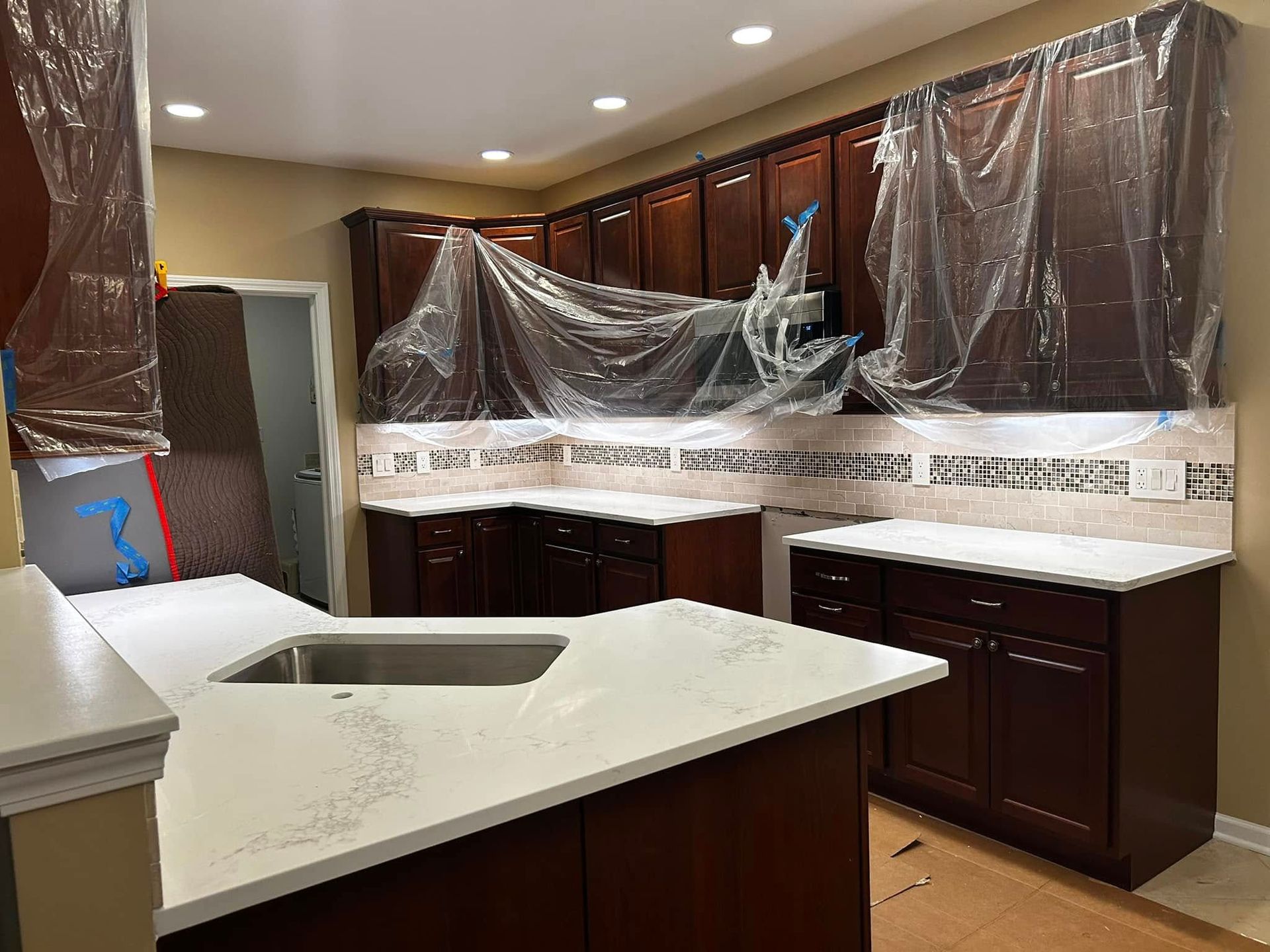 Kitchen under renovation with dark wood cabinets, white countertops, and plastic coverings over surfaces