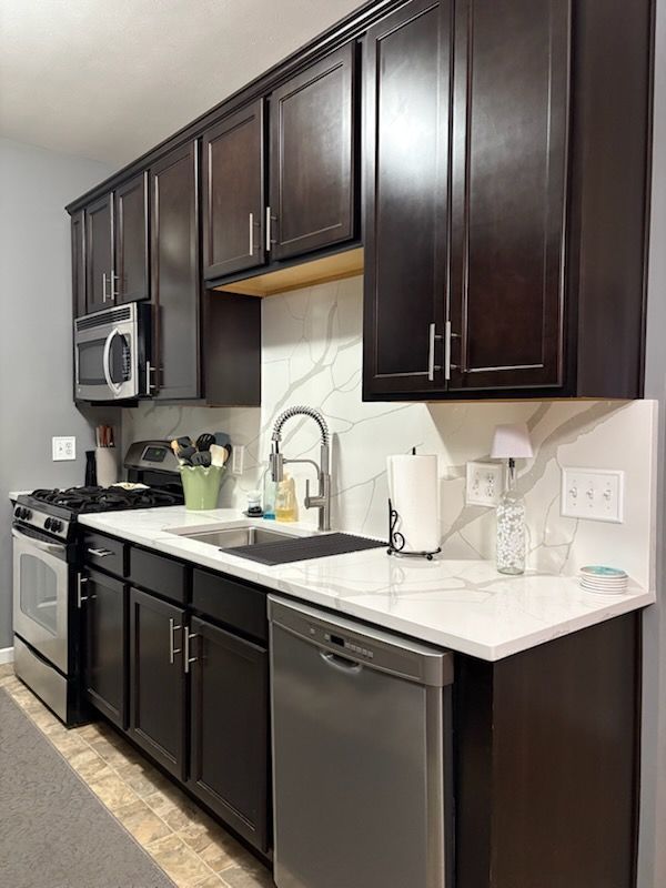 Dark wood kitchen cabinets with white countertops, stainless appliances, and a sink along a narrow galley wall.