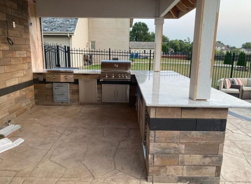 Covered outdoor kitchen with stone counters, built-in grill, and tiled floor on a patio.