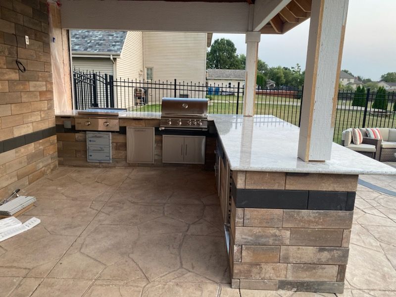 Covered outdoor patio with stone counter, built-in grill, and tiled floor beside a fenced yard.