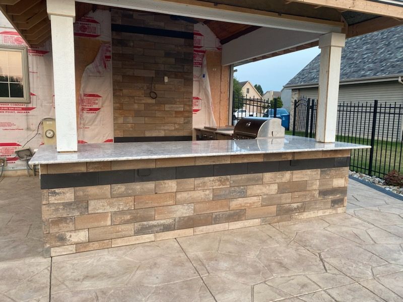 Covered outdoor patio with stone half-wall, tiled floor, and brick fireplace under construction