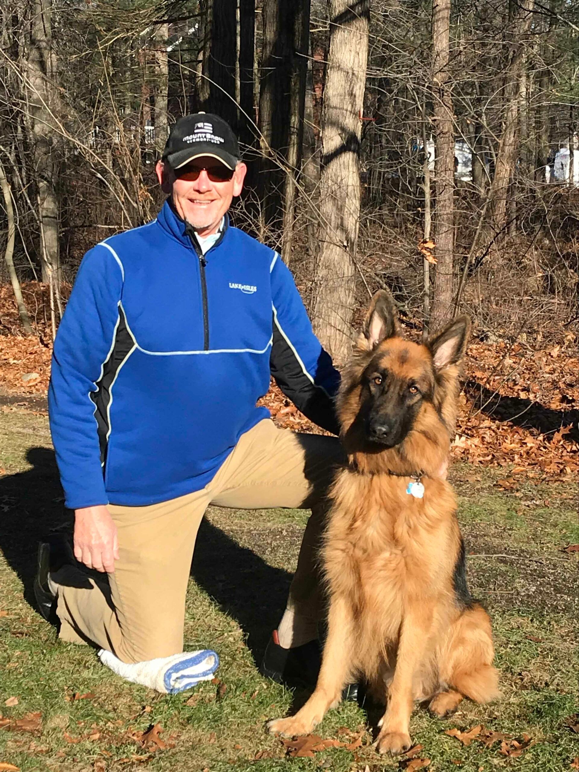 Man kneeling beside a German Shepherd dog outdoors. Man wears blue top, tan pants. Dog is tan and black.