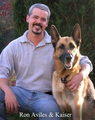 Man with graying hair, smiling, seated with German Shepherd dog, both looking toward viewer. Outdoor setting.