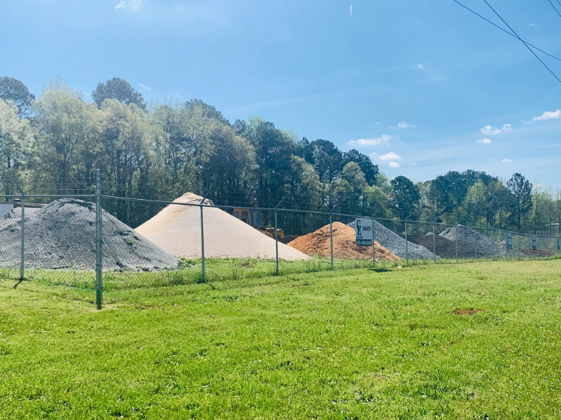 Gravel and sand piles in a fenced grassy field with trees and blue sky in the background
