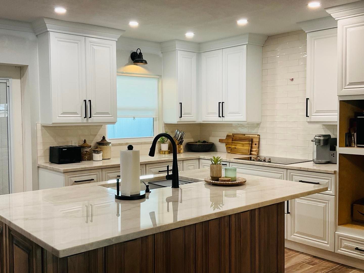 A bright, modern kitchen featuring white cabinets, a light stone island with a dark wood base, and a black faucet.