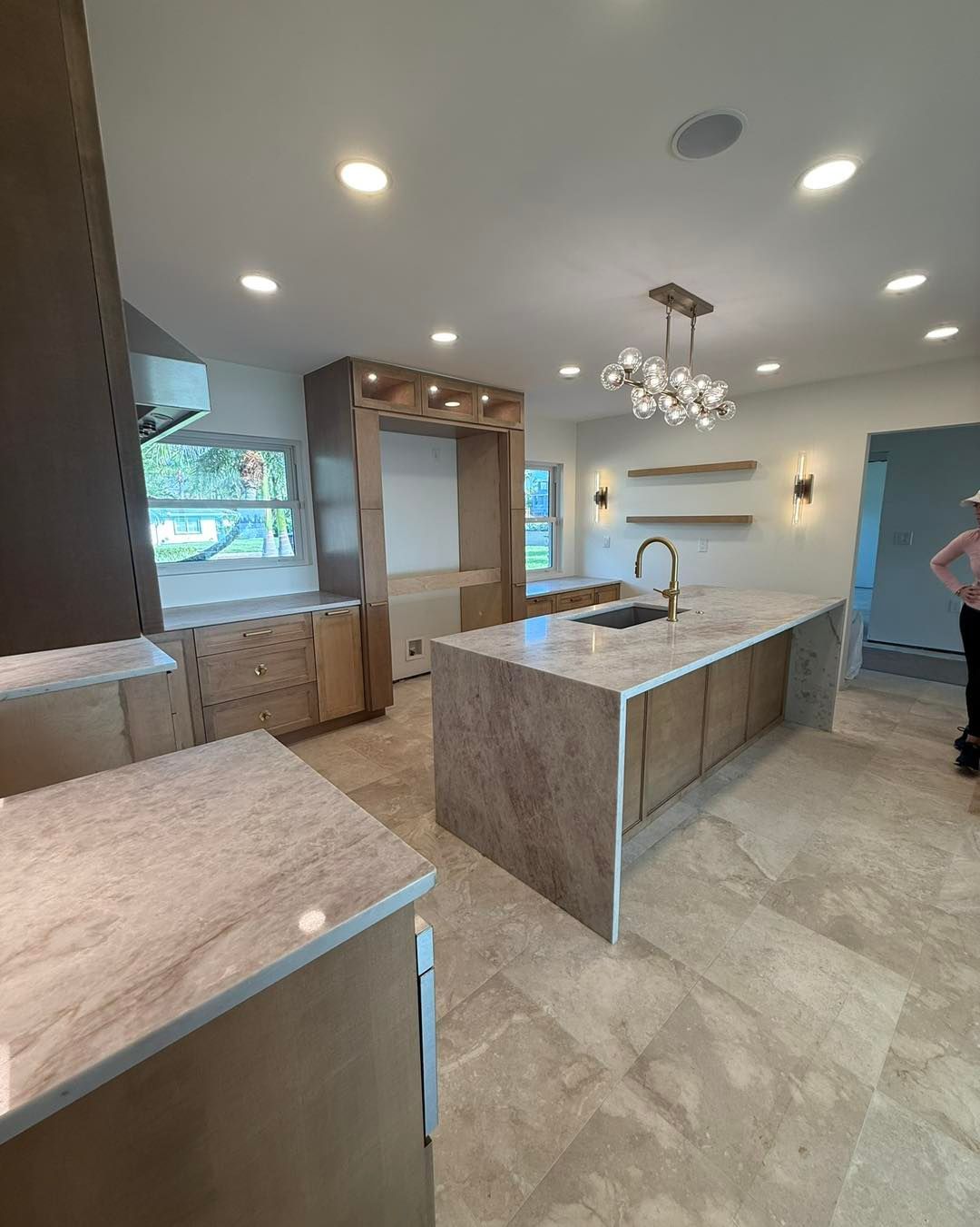 A kitchen featuring a large island with a light granite countertop, wood-toned cabinetry, tile flooring, and a chandelier.