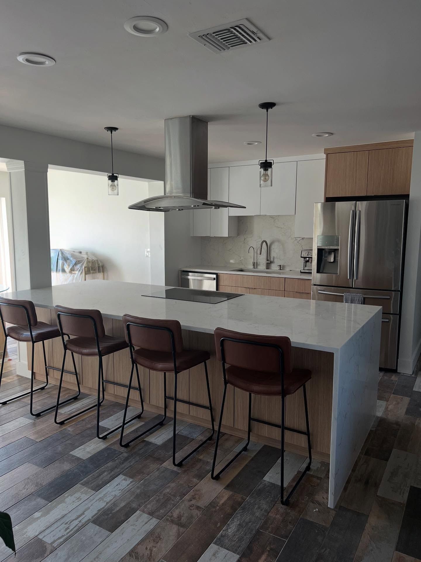 Modern kitchen with a long white island, four brown stools, stainless steel appliances, and wood-toned cabinets.