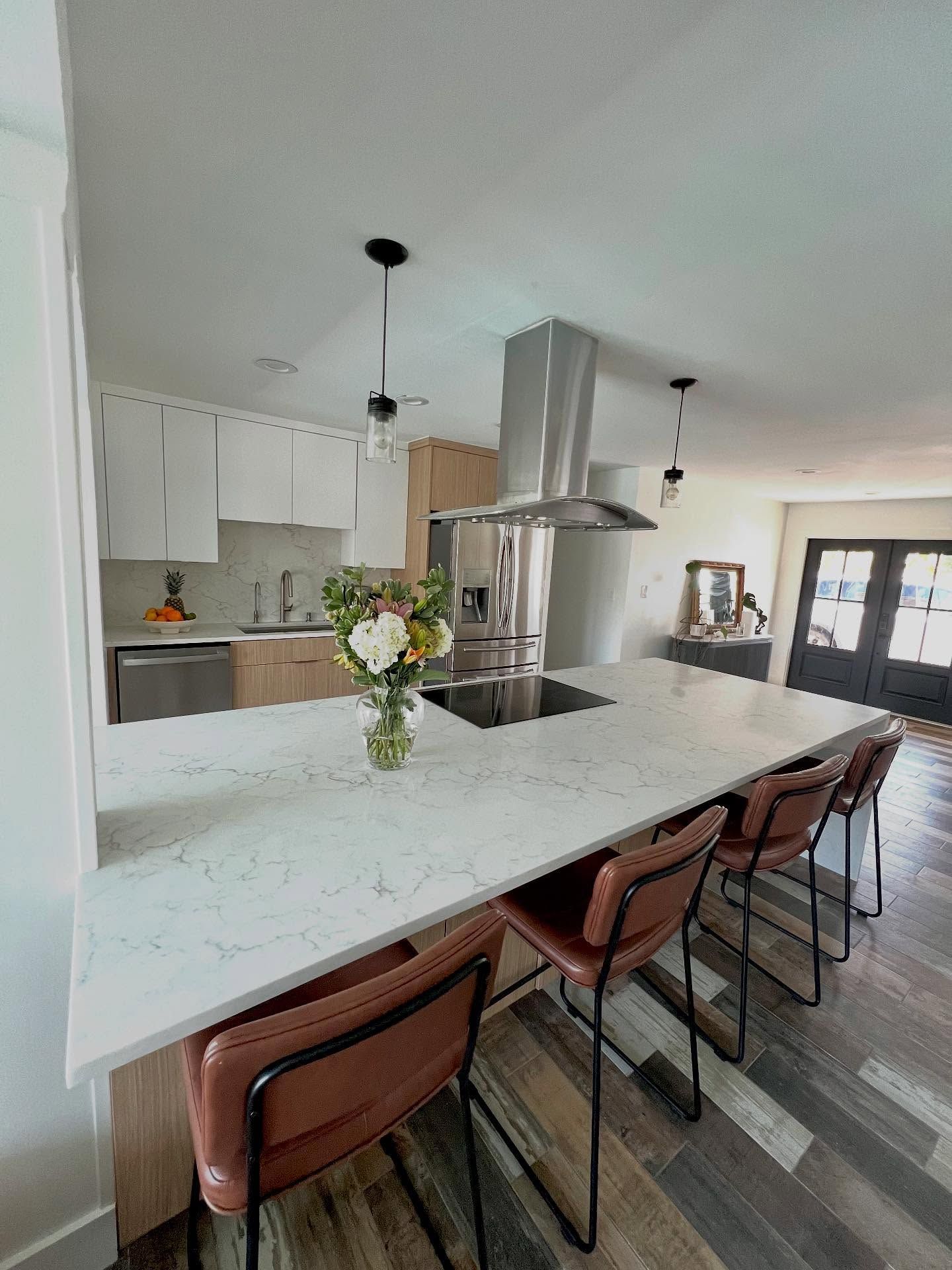 A kitchen with a large white marble island, three leather stools, white cabinetry, and stainless steel appliances.