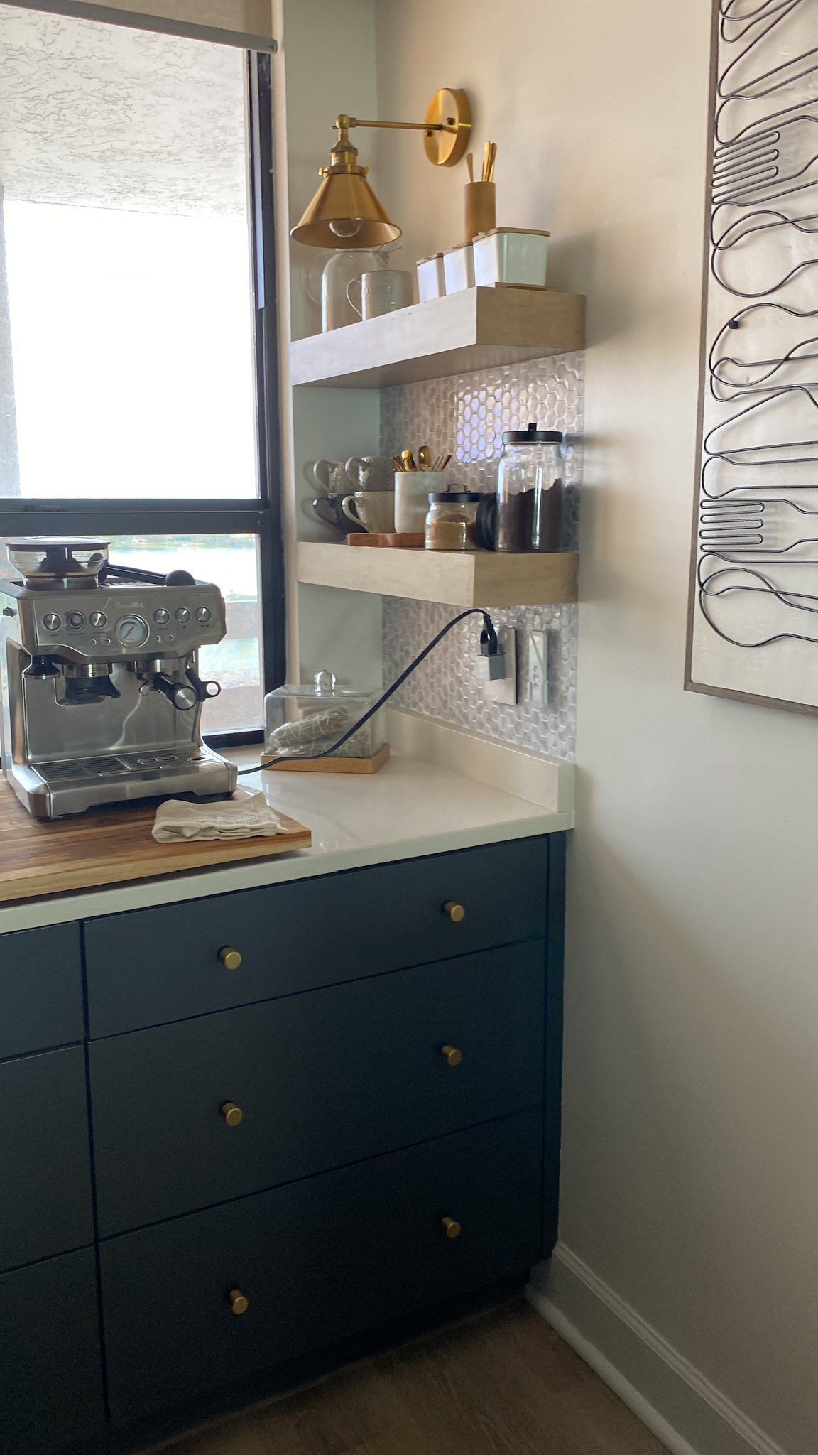 A navy blue coffee station with a silver espresso machine, two light wood shelves, and a decorative wall hanging.