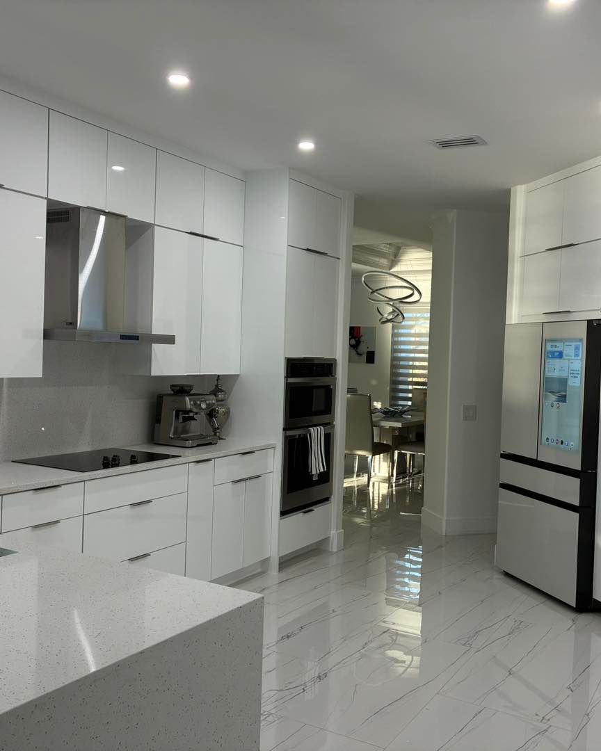 A modern white kitchen featuring glossy cabinets, a stainless steel range hood, built-in ovens, and marble-look tile floors.