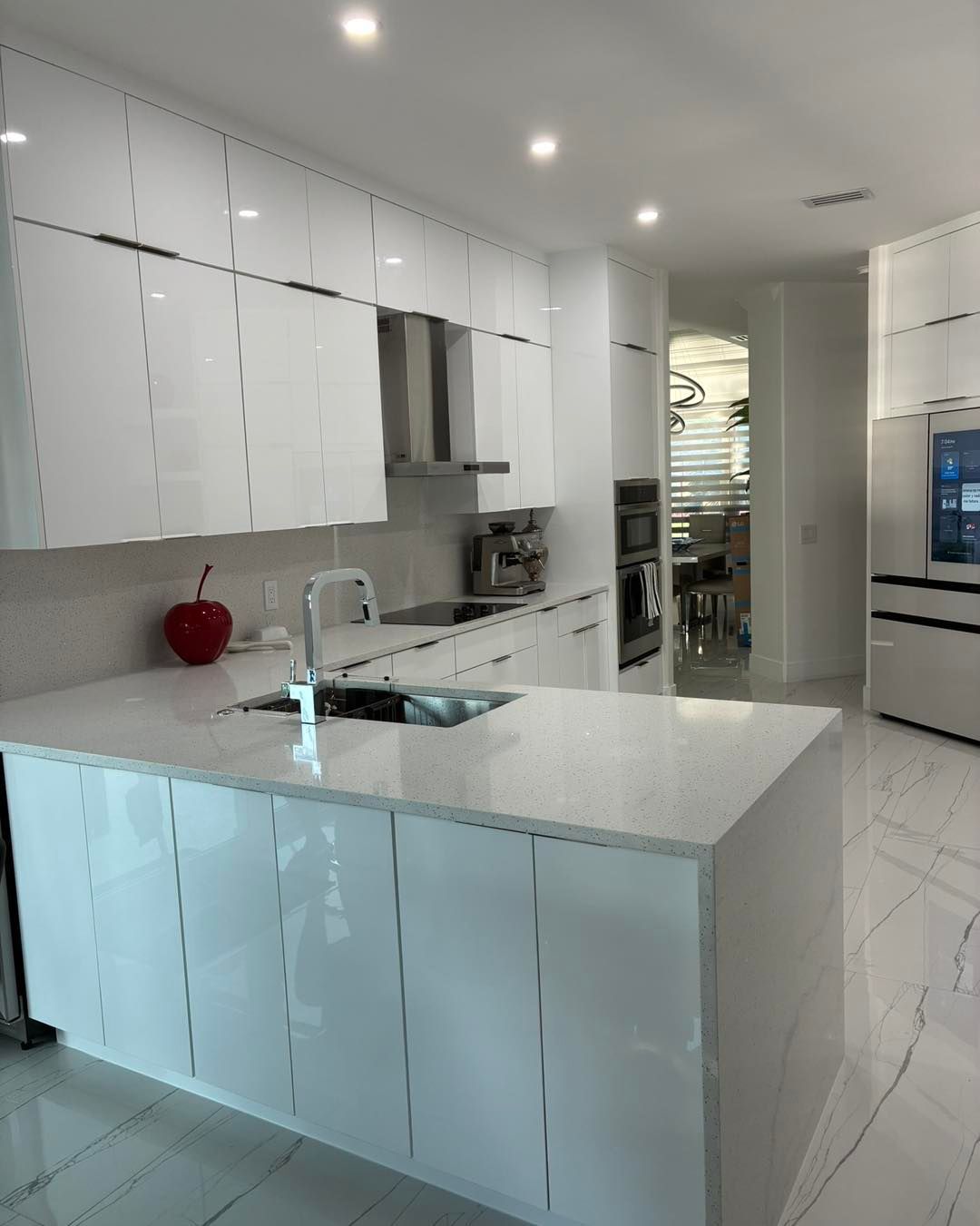 A modern, white high-gloss kitchen with a stone island countertop, silver sink fixture, and marble-style floor tiles.