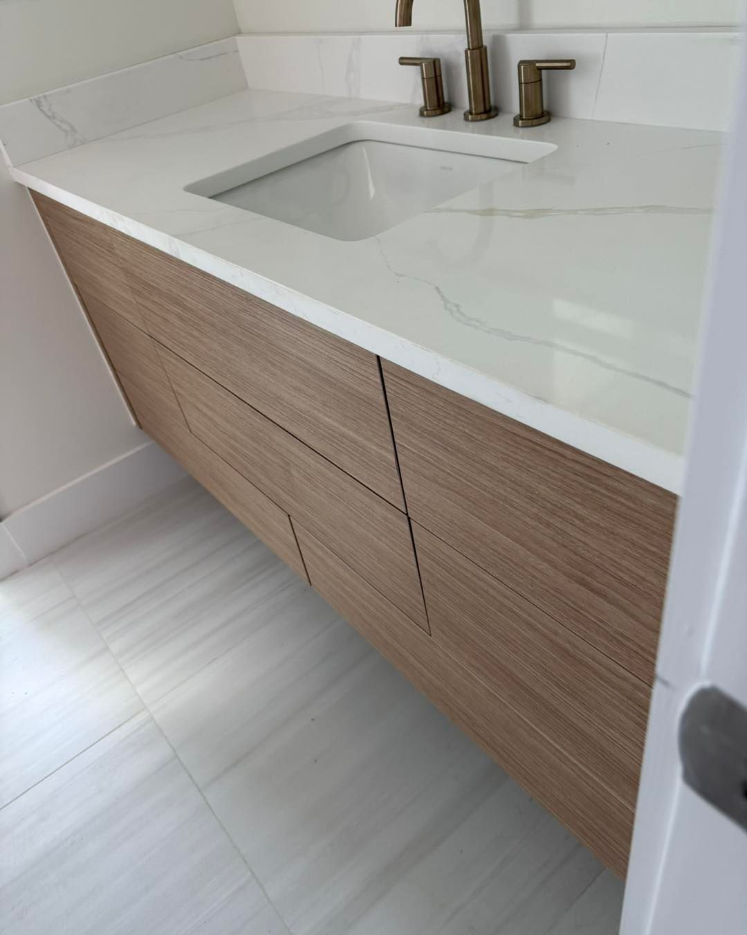 Modern wood-grain vanity with a white marble countertop and gold faucet in a bathroom with light tile flooring.