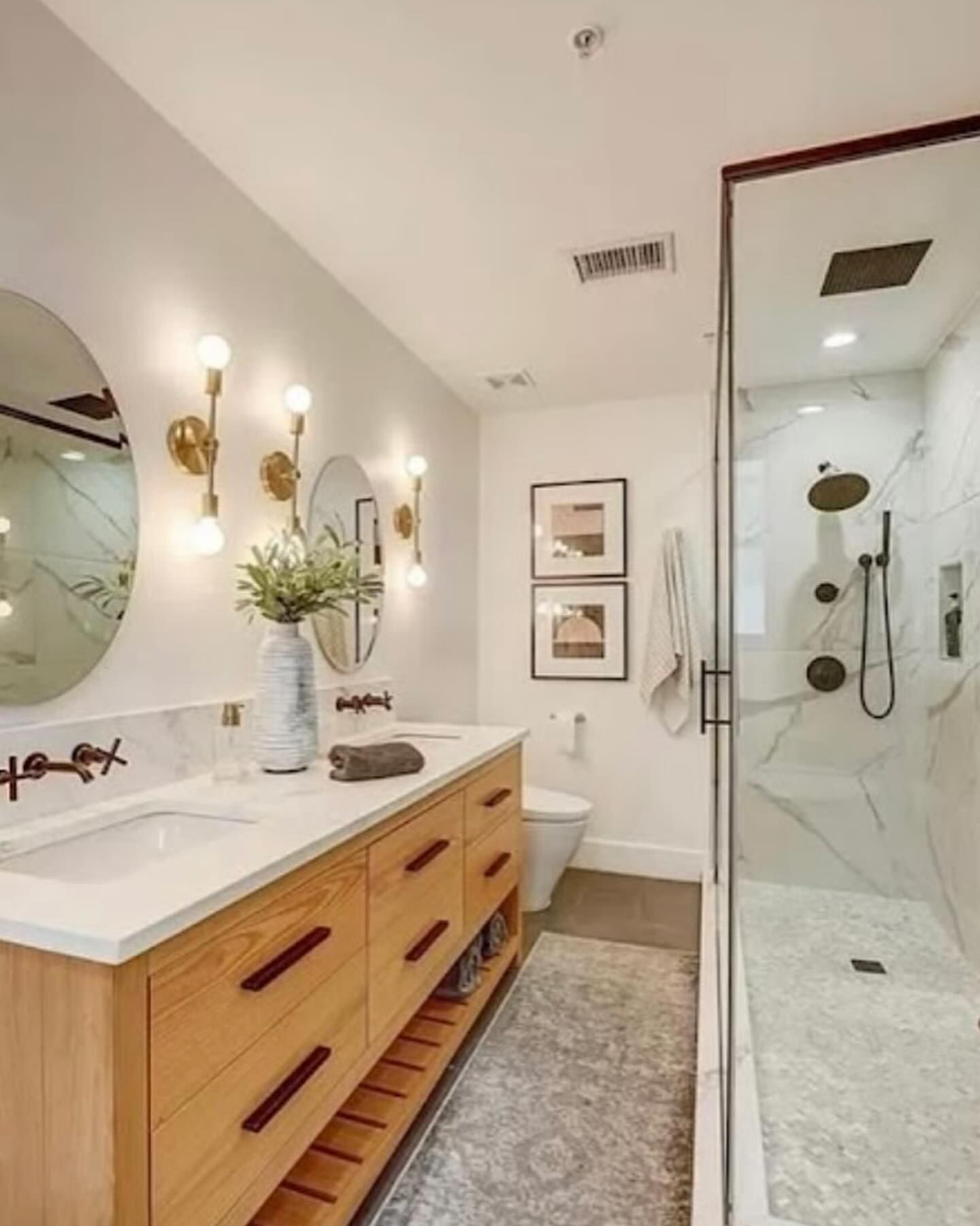 A bright, modern bathroom with a light wood vanity, white countertops, dual circular mirrors, and a glass-enclosed shower.